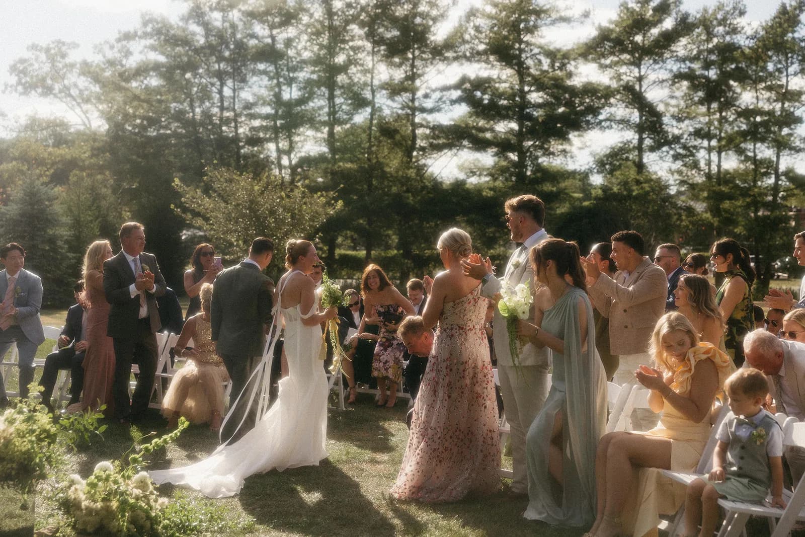 Couple walk the aisle between applauding guests, a maid of honor holding a small white and green bouquet