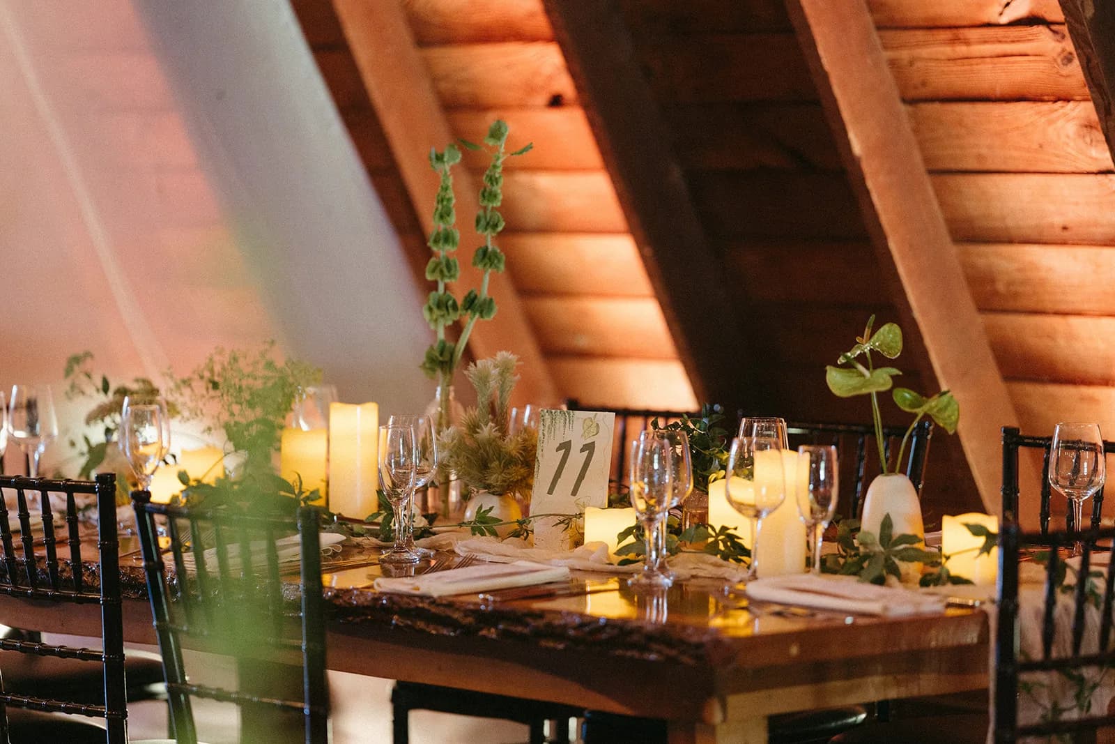 Reception table under timber beams with bells of Ireland, Phylica, and green anthurium in bud vases beside pillar candles