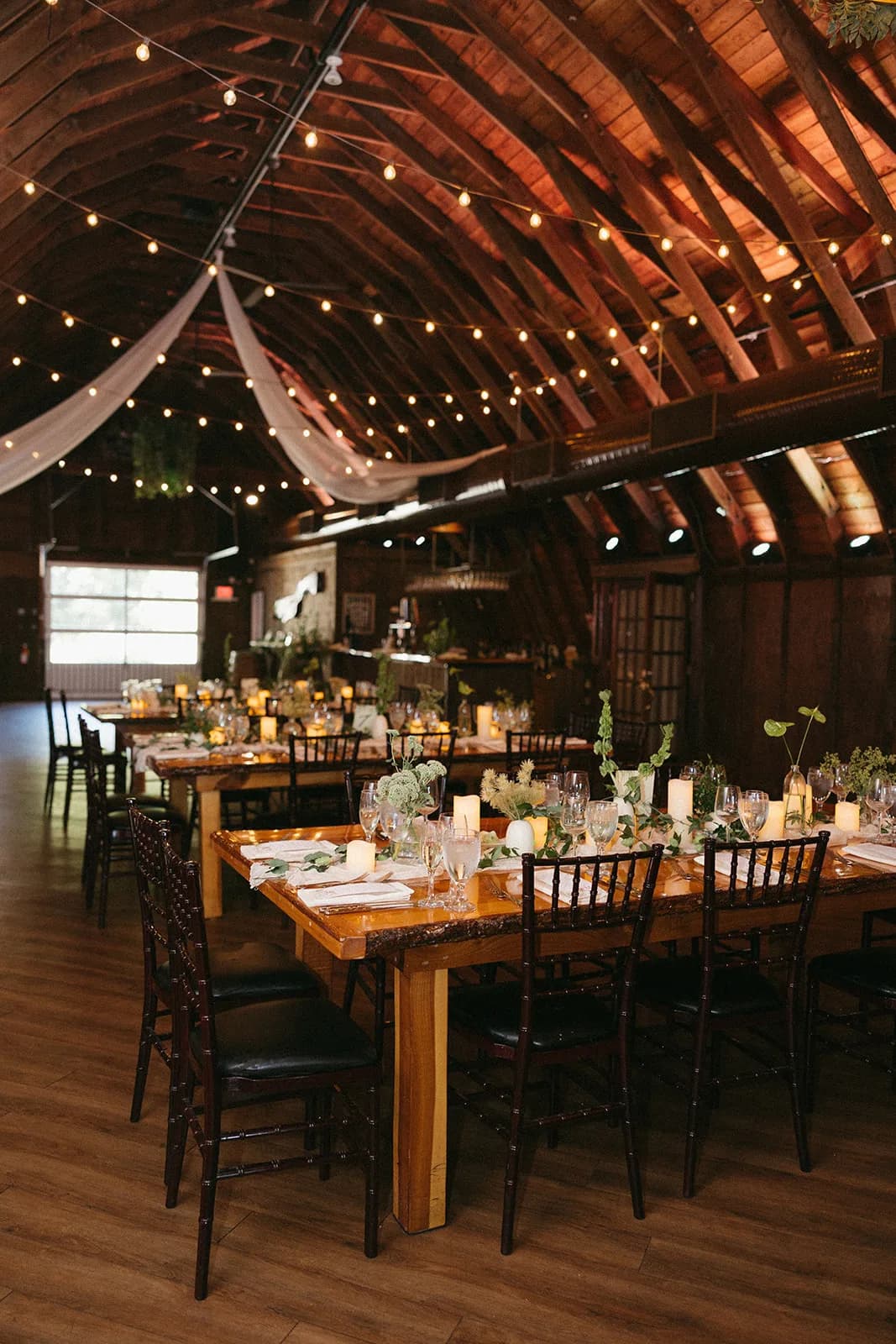 Barn reception with string lights and draped fabric, long wooden tables set with candles and green bud vase arrangements