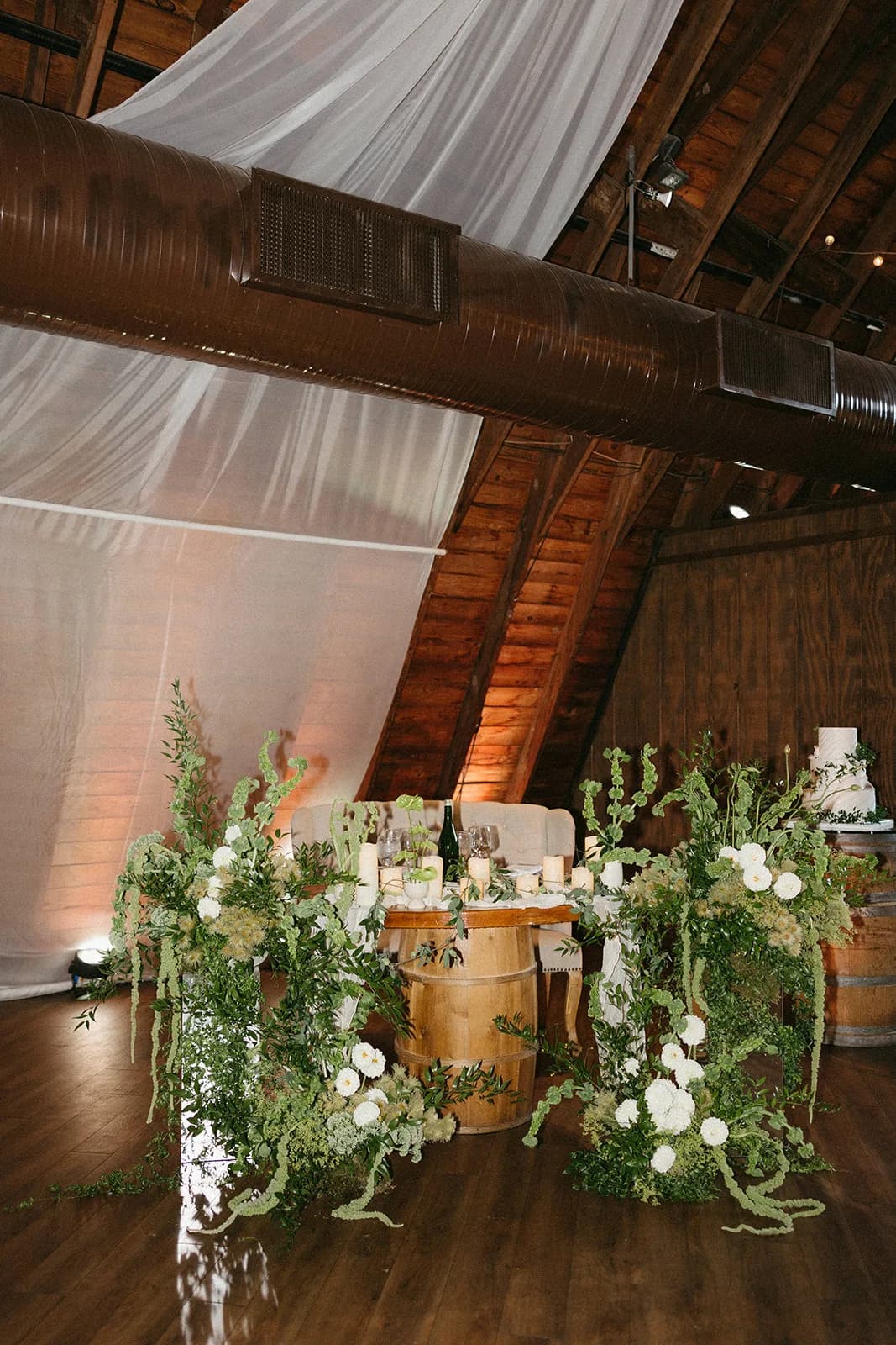 Sweetheart table on a wine barrel between tall deconstructed arrangements of white dahlias, bells of Ireland, and amaranthus