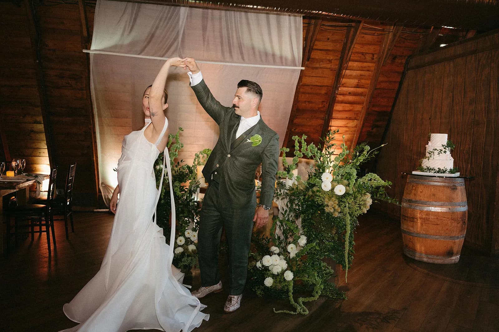 Groom twirls the bride during the first dance beside sculptural arrangements of white dahlias and trailing green amaranthus