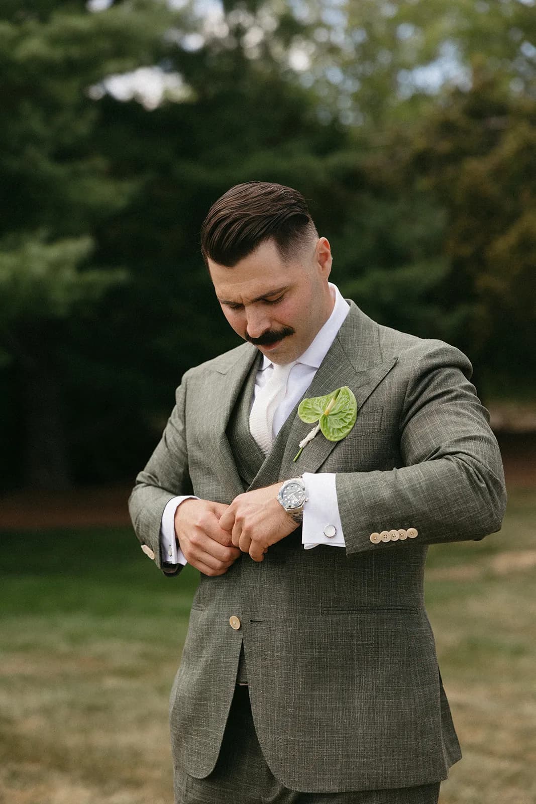 Groom in a gray green suit fastens his cuff, a single green anthurium boutonniere pinned to his lapel