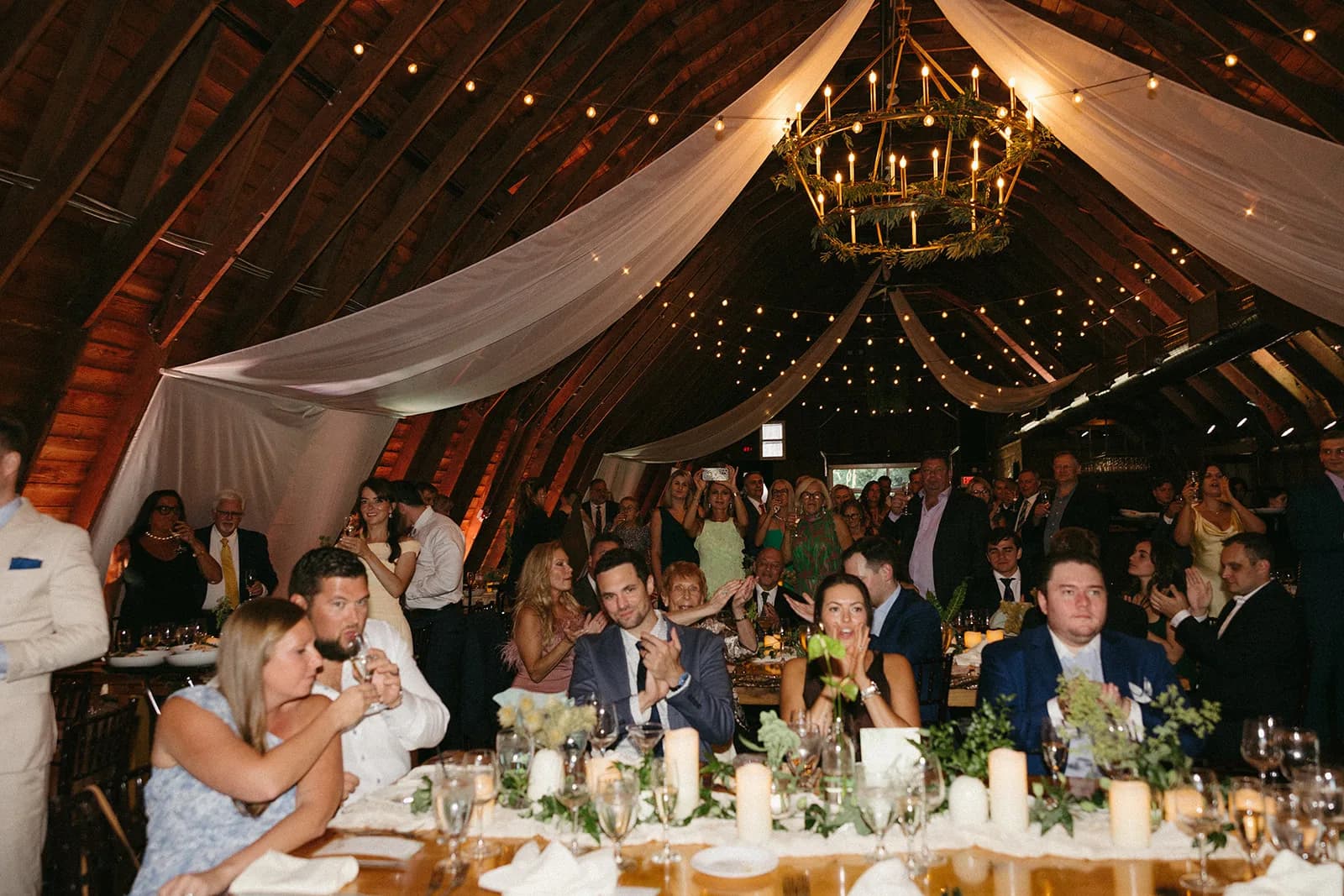 Guests applaud in the barn beneath a greenery-wrapped candle chandelier, a single anthurium in a bud vase on the table