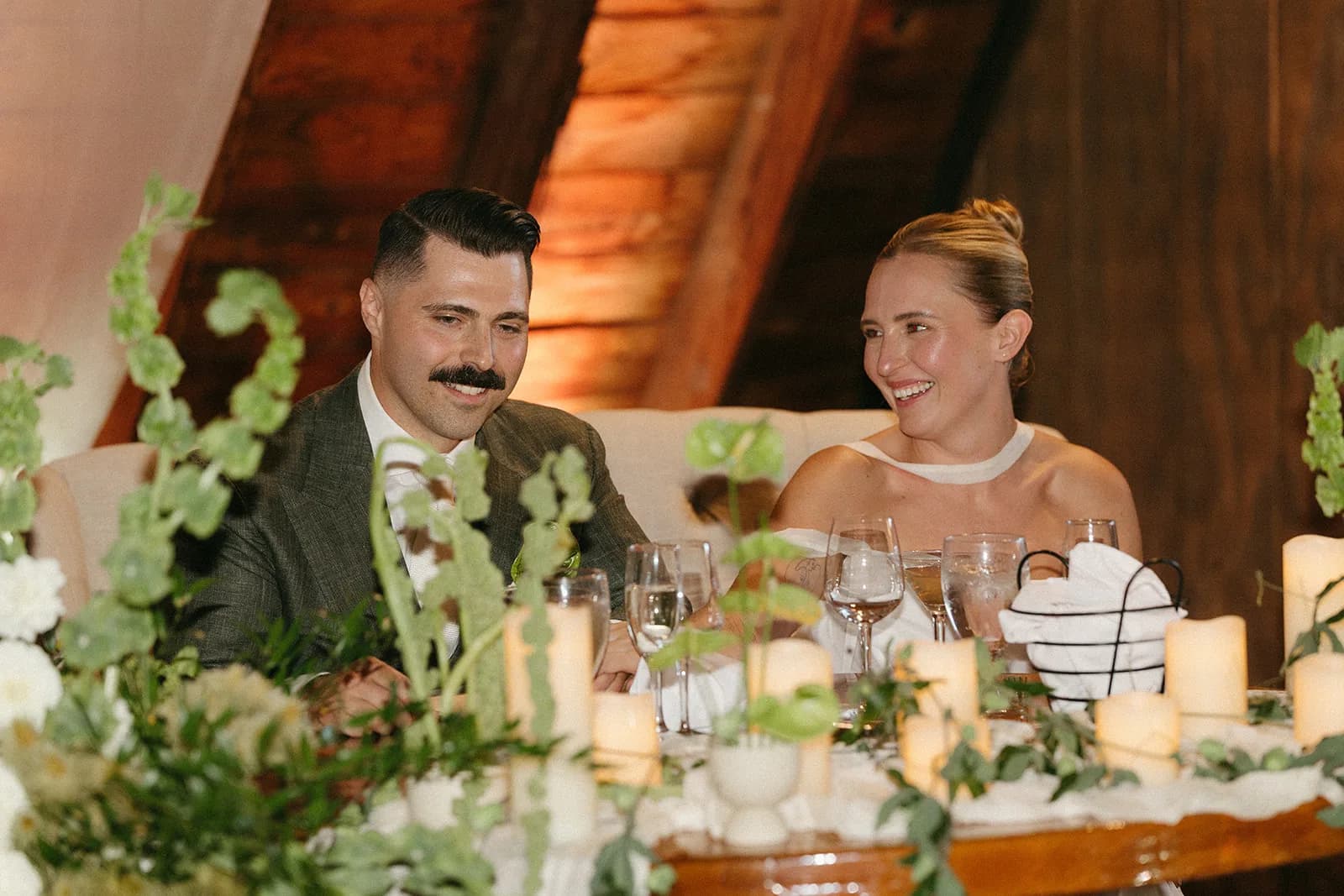 Couple laugh at the sweetheart table behind bells of Ireland stems, white dahlias, and a row of pillar candles