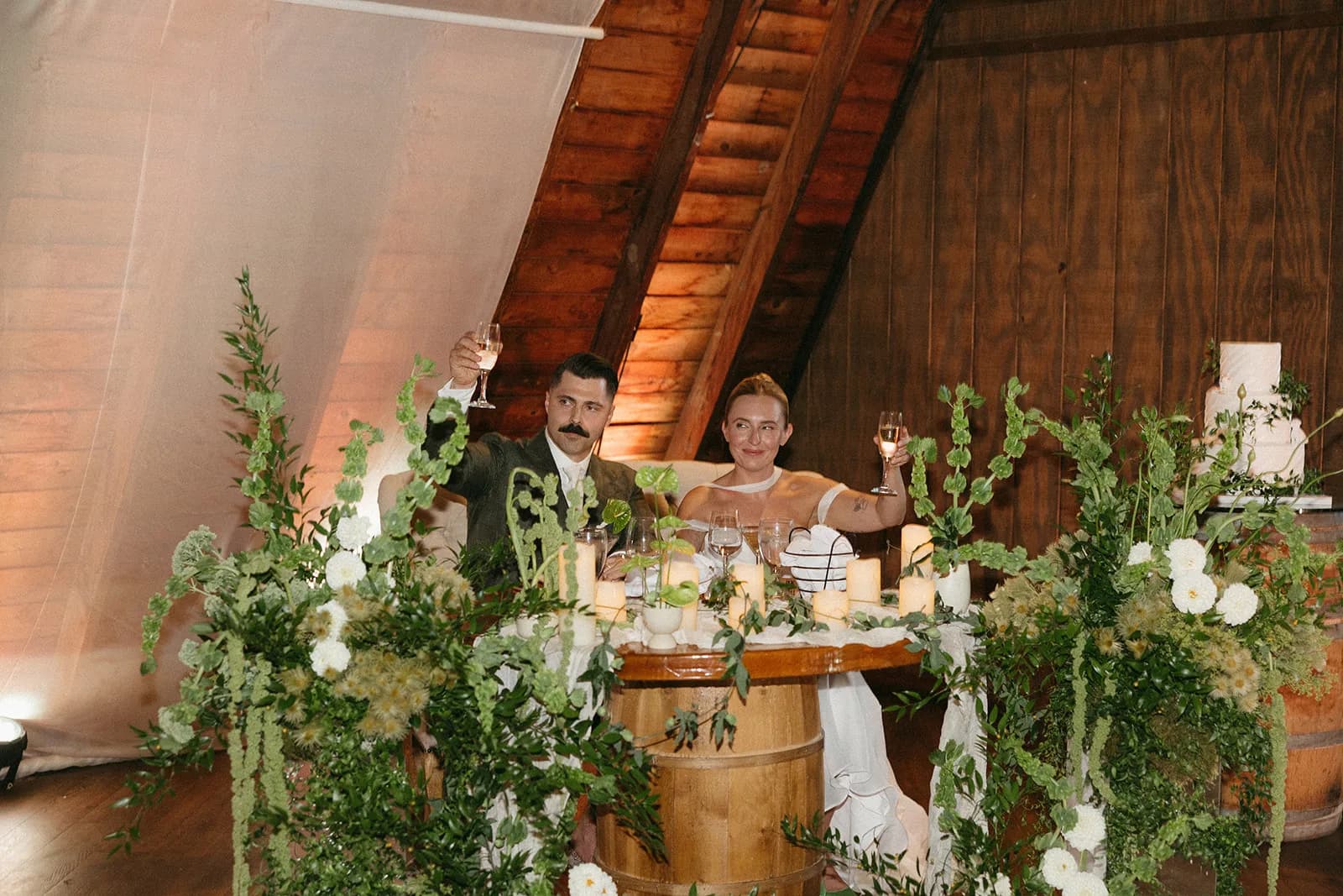 Couple raise glasses at the sweetheart table between tall arrangements of bells of Ireland, white dahlias, and amaranthus