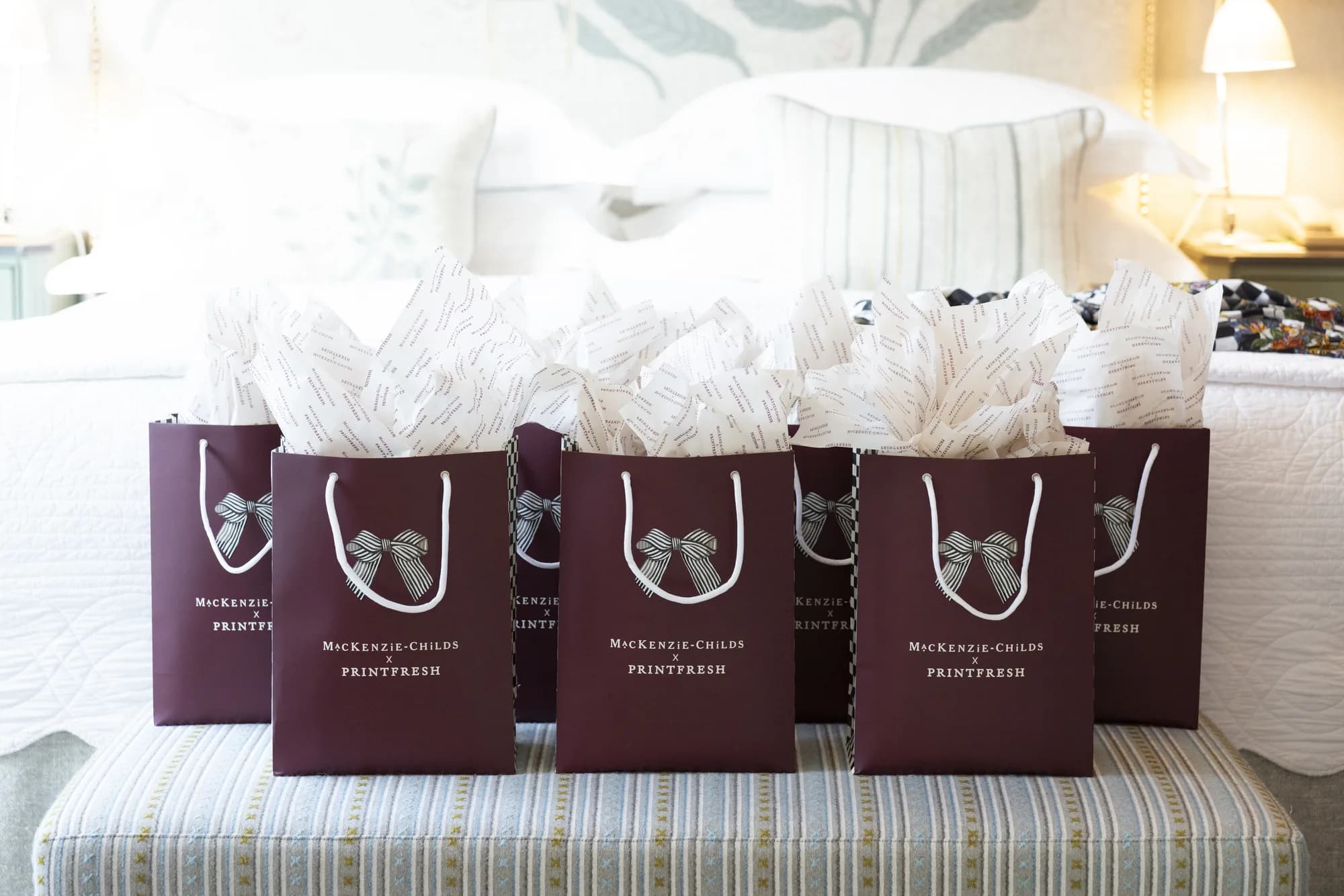 Six burgundy MacKenzie-Childs x Printfresh gift bags with striped bows sit on a bench at the foot of a hotel bed