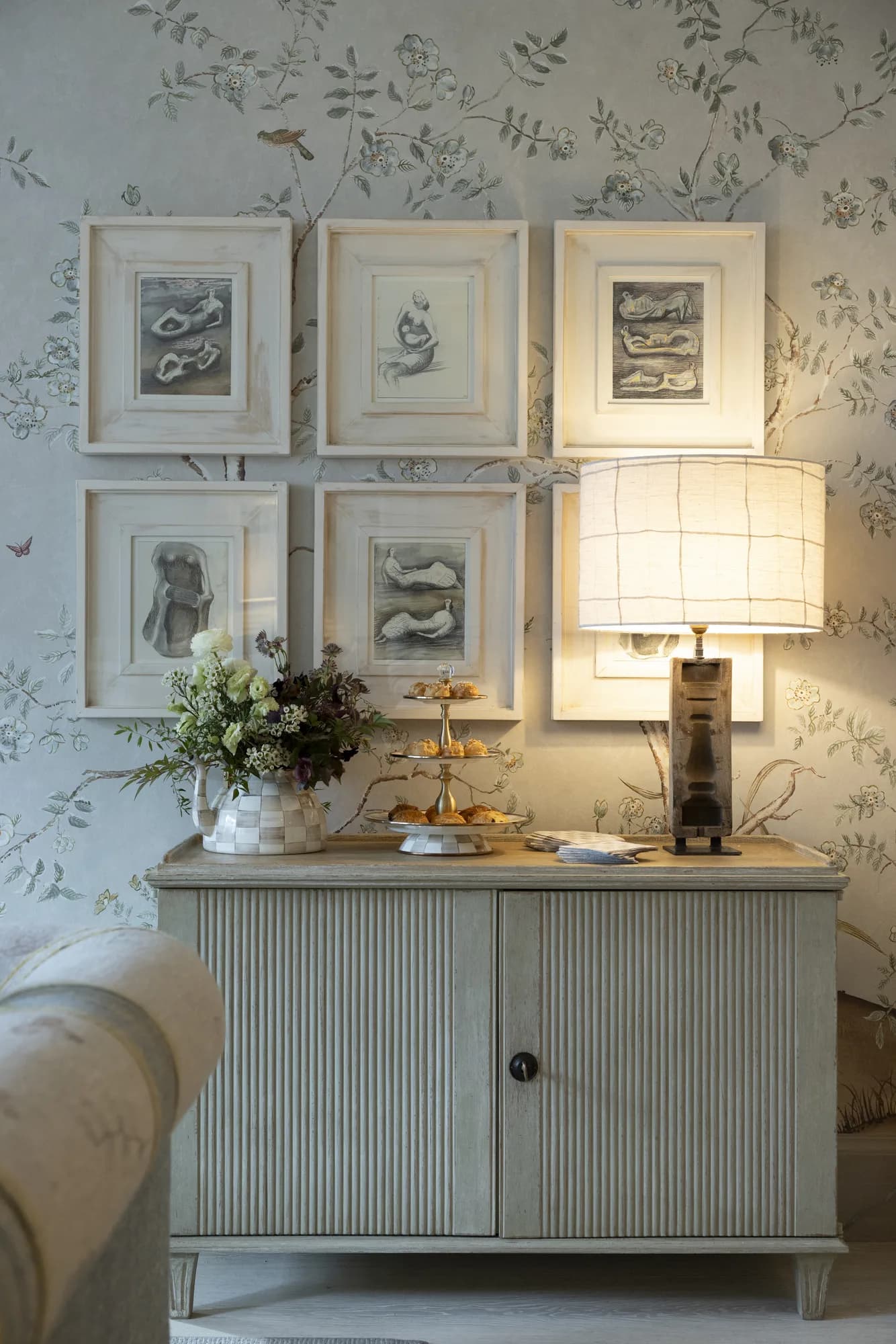 A small seasonal arrangement and tiered pastries on a fluted sideboard beneath framed sketches and floral wallpaper