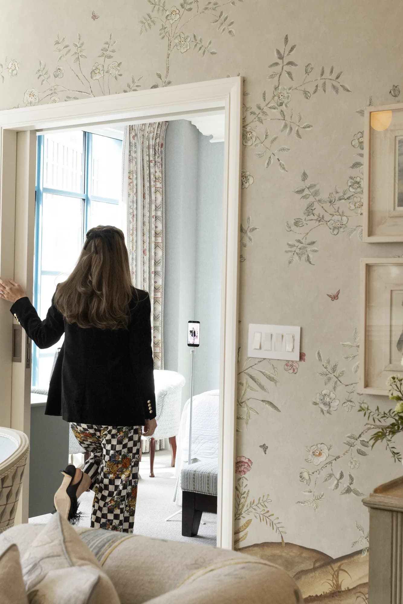 A guest in a black blazer and checkered floral pajama pants passes through a doorway in a wallpapered suite
