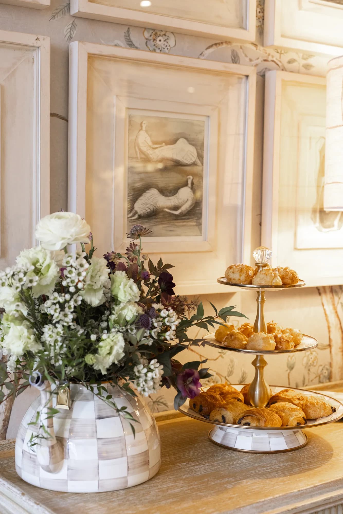 White ranunculus, waxflower and plum hellebore in a MacKenzie-Childs check vase beside a tiered stand of pastries