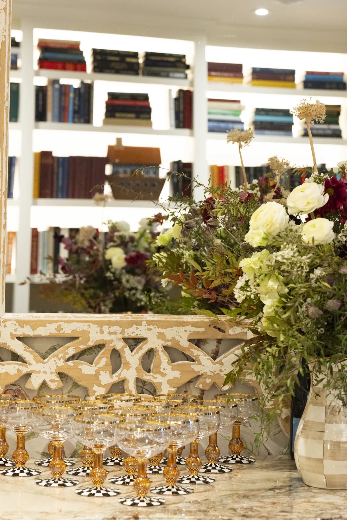 Amber coupes on check bases line a marble bar beside a vase of ranunculus, cosmos and tall dried allium seedheads