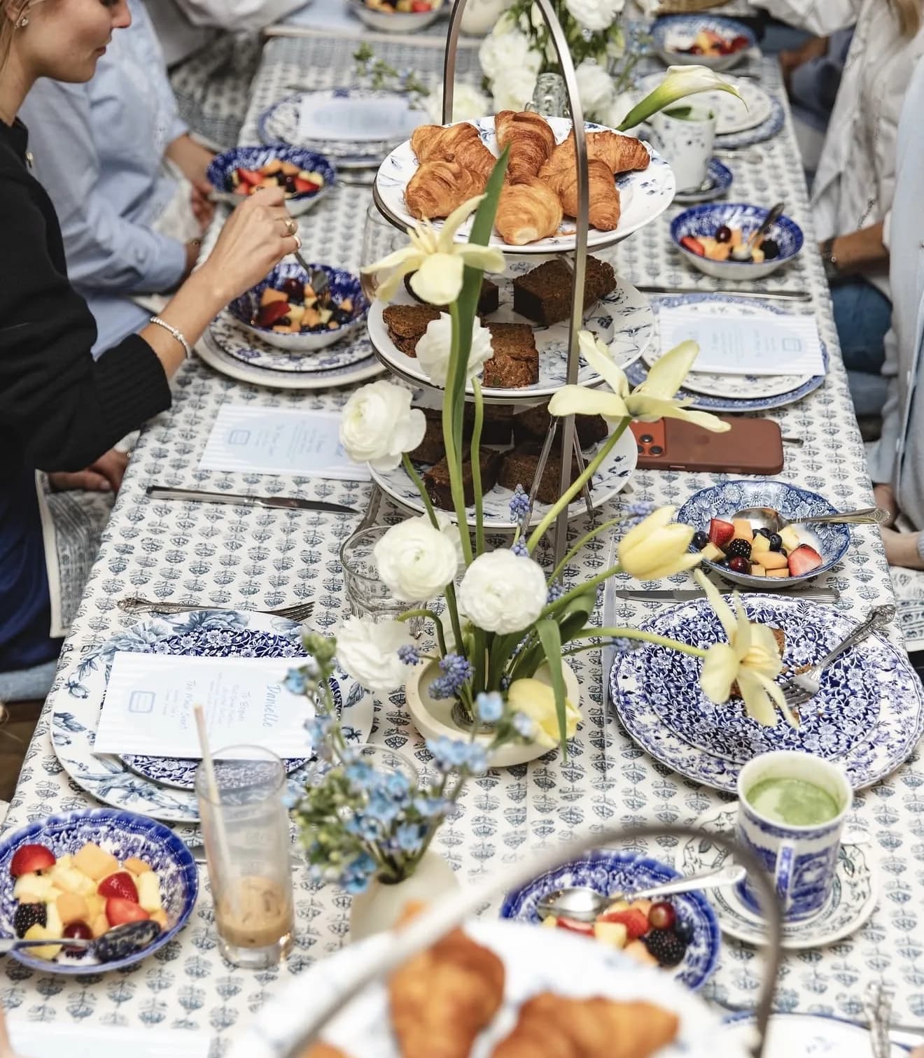 Guests along a blue and white block print table with white ranunculus, yellow tulips and muscari beside a tiered pastry stand