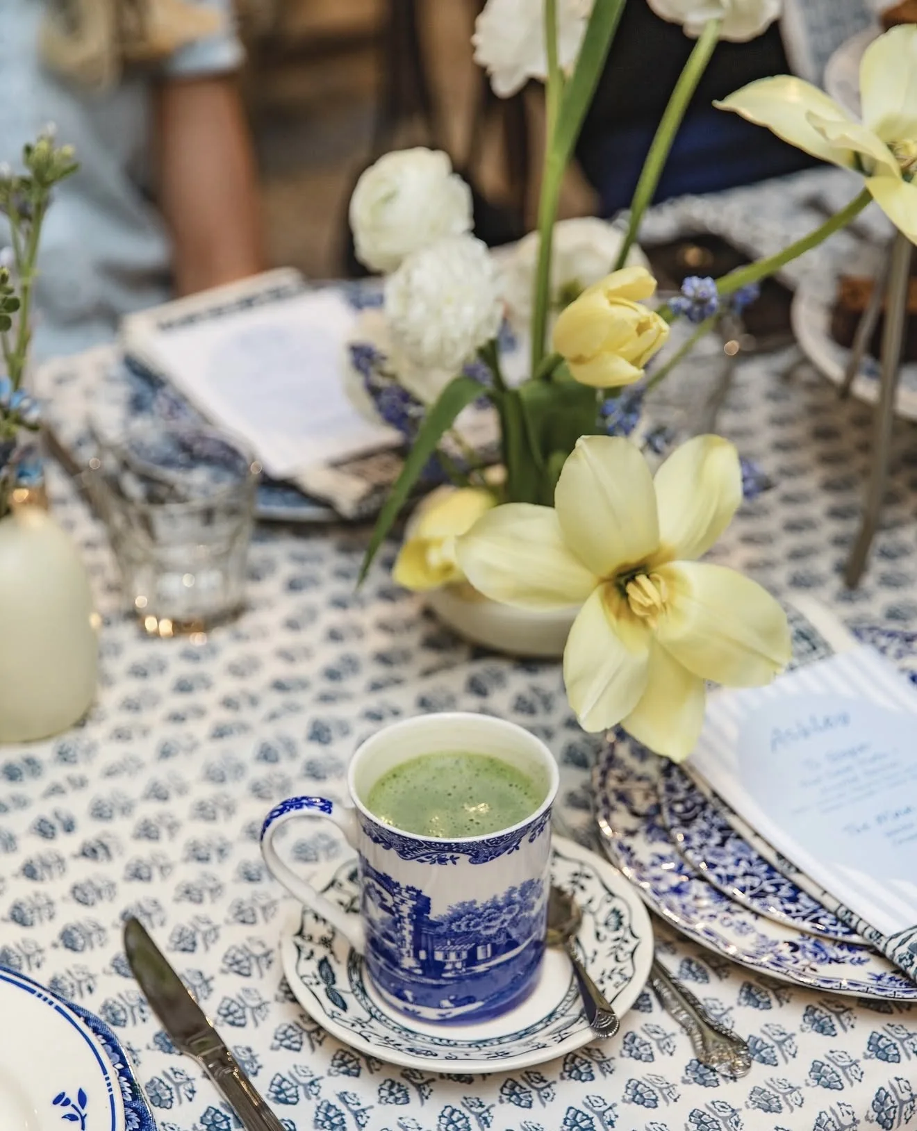 Open yellow tulip, white ranunculus and muscari in a small white vase beside a blue transferware mug of matcha and a menu