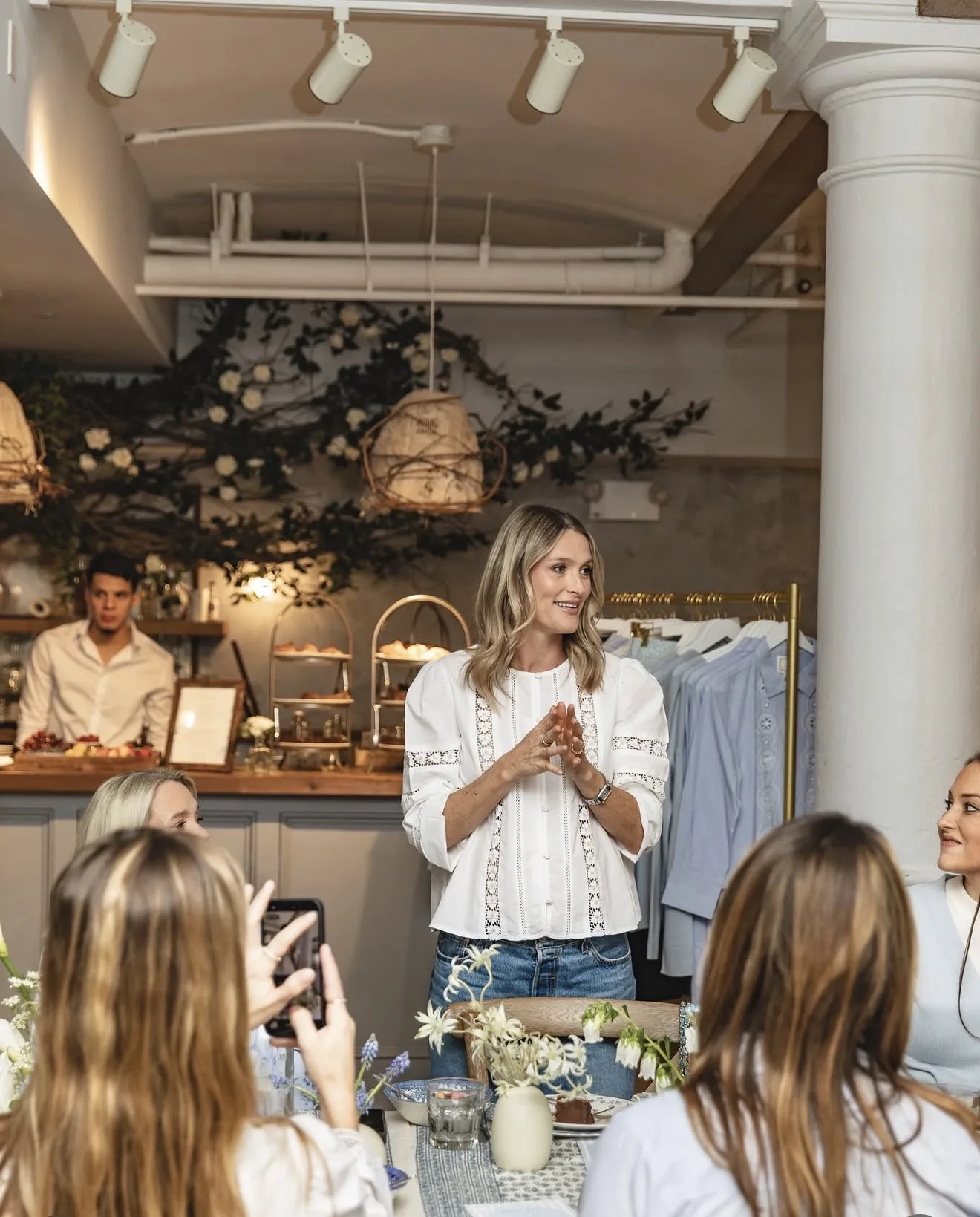 Host speaks at the head of the table beneath a vine and white rose canopy, with a rack of blue Sail to Sable shirts behind