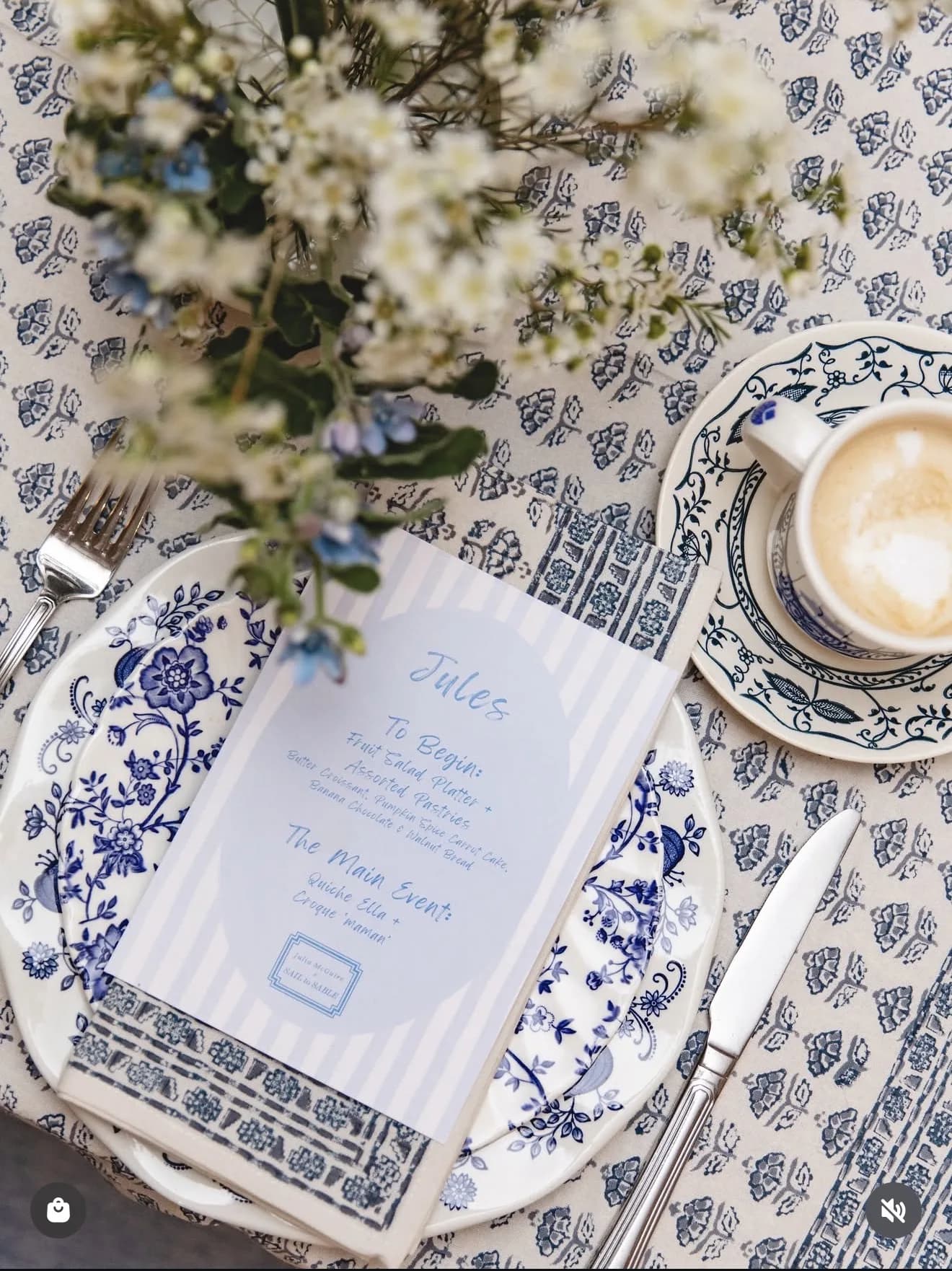Waxflower and forget-me-not stems trail across a blue and white place setting with a striped menu card and a cappuccino