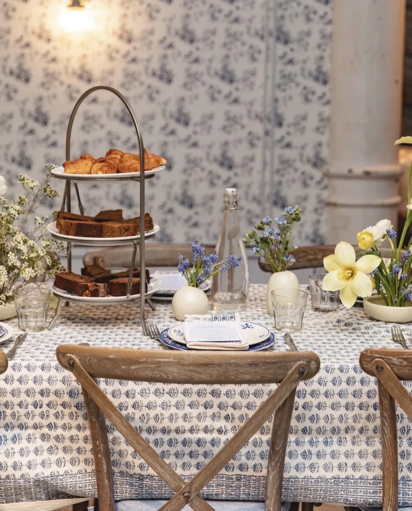 Tiered pastry stand and bud vases of muscari, forget-me-nots and a yellow tulip on block print cloth against blue toile paper