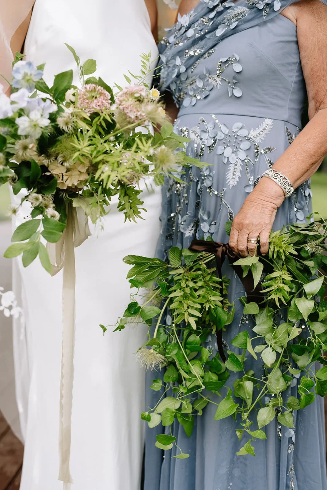 Bride holds a bouquet of pale blue nigella, blush scabiosa and hydrangea beside a trailing greenery bouquet in brown ribbon