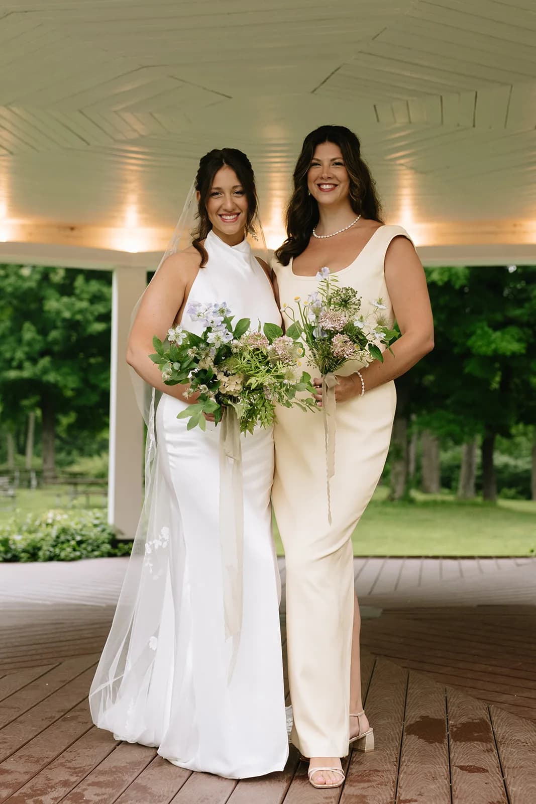 Shayna and an attendant in cream beneath a pavilion ceiling, each holding a loose bouquet of nigella, scabiosa and greenery