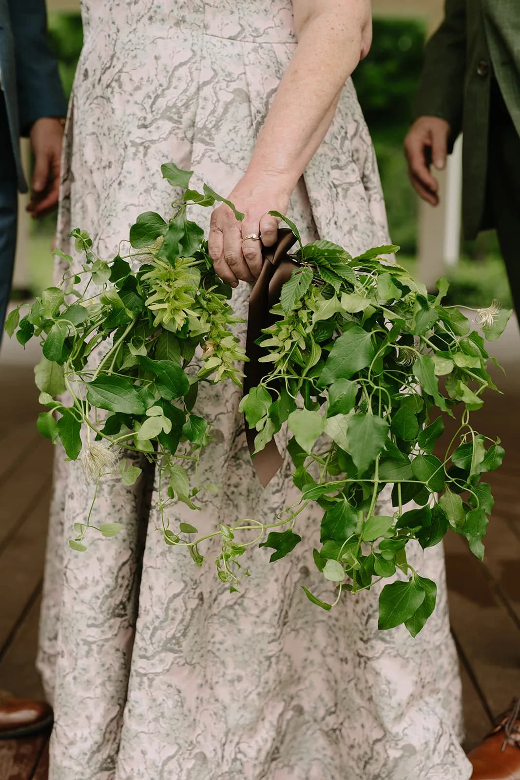 Woman in a pale floral gown holds a trailing greenery bouquet of ivy, seeded vines and clematis seed heads with brown ribbon