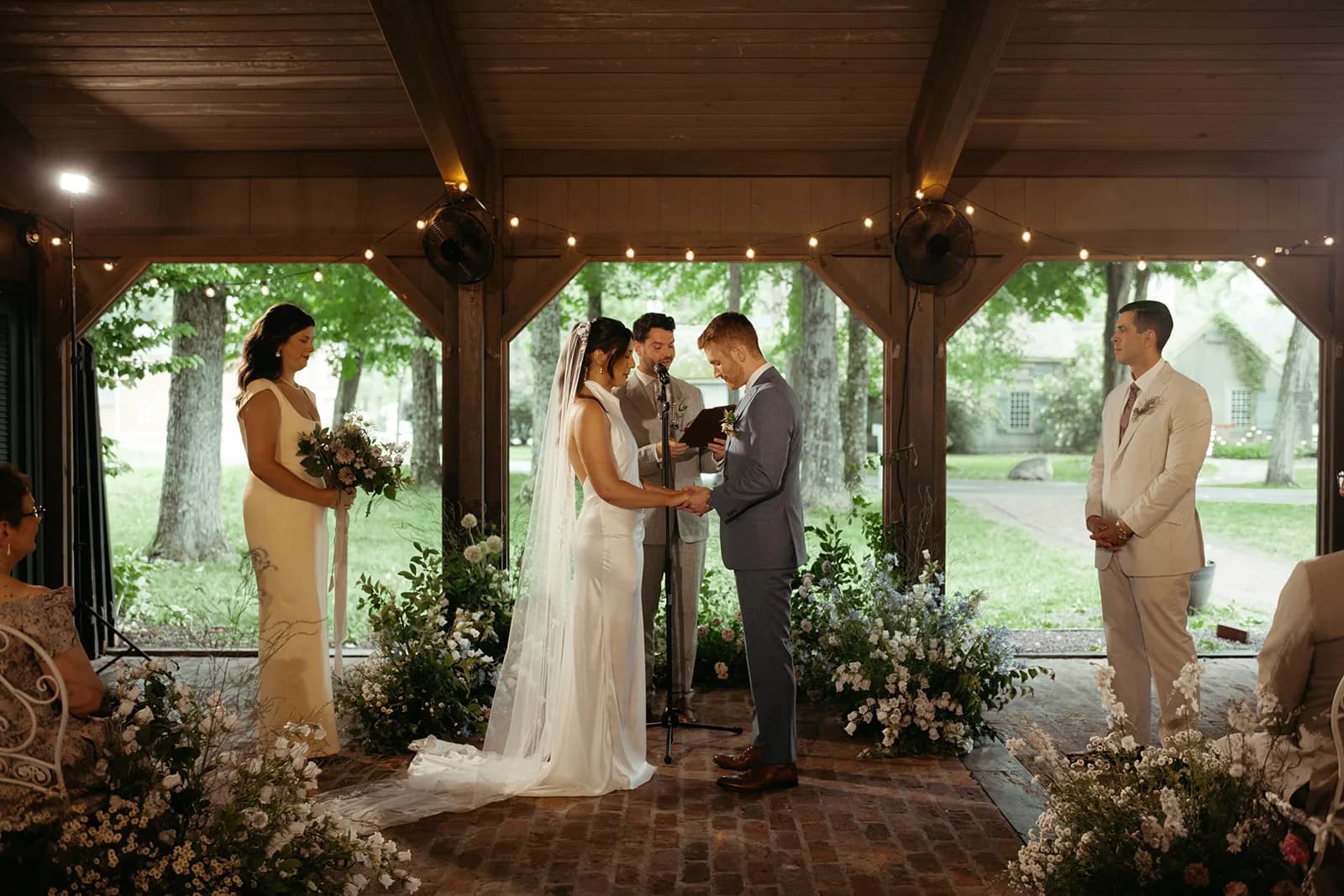 Shayna and Evan hold hands at the microphone beneath a timber pavilion, a crescent of white and blue blooms at their feet