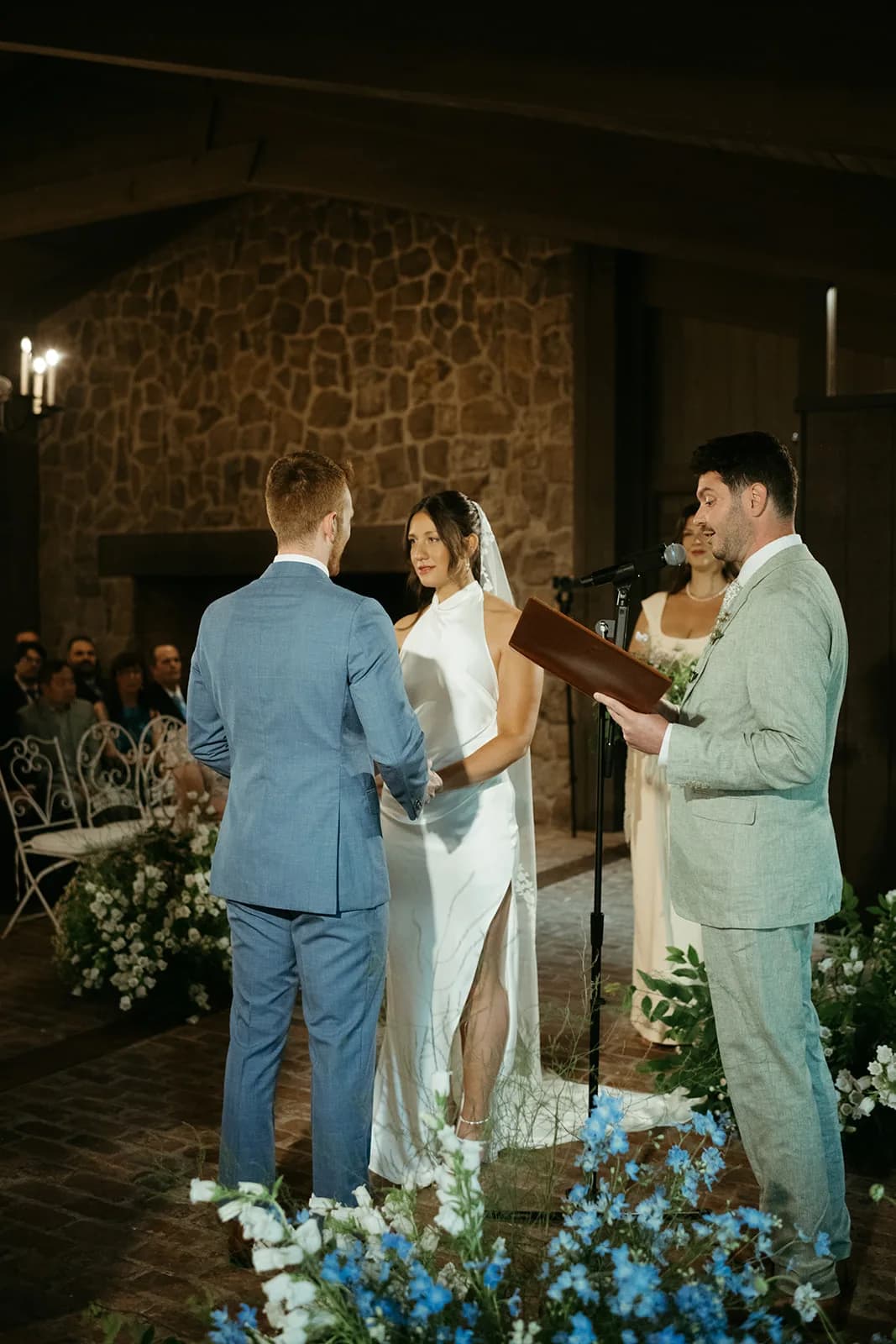 The couple face each other during vows before a stone fireplace, blue delphinium and white campanula rising in the foreground