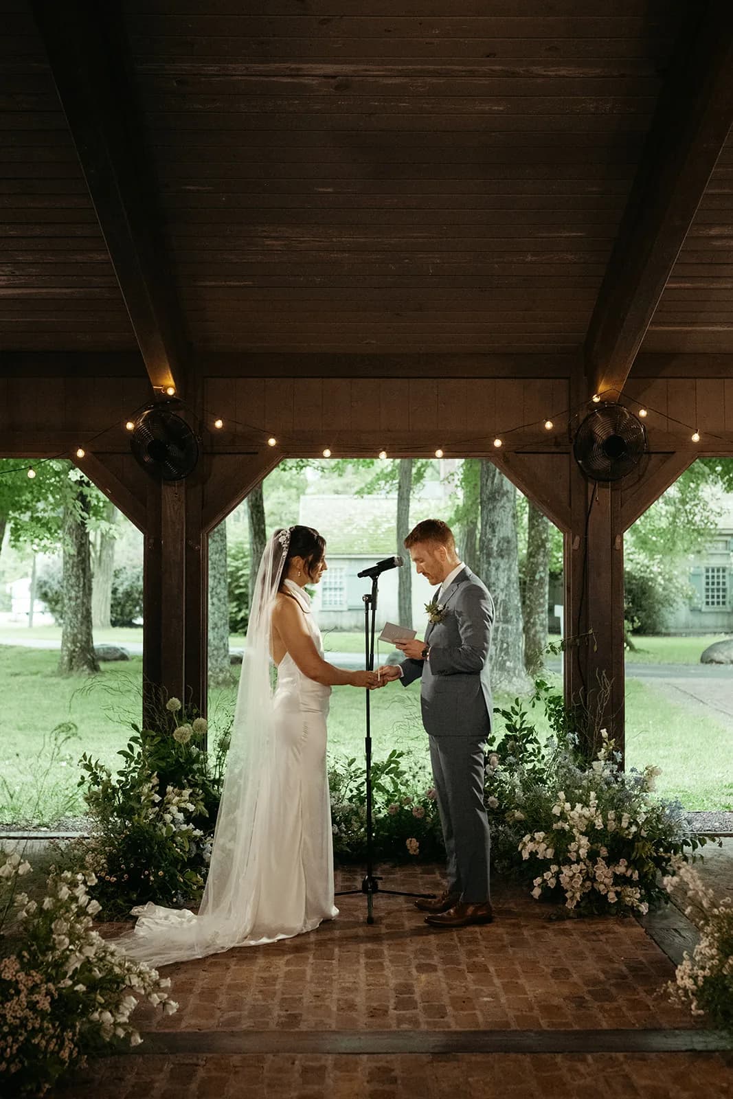 Evan reads vows to Shayna at the microphone, her veil trailing, white campanula and allium clustered at the pavilion edge