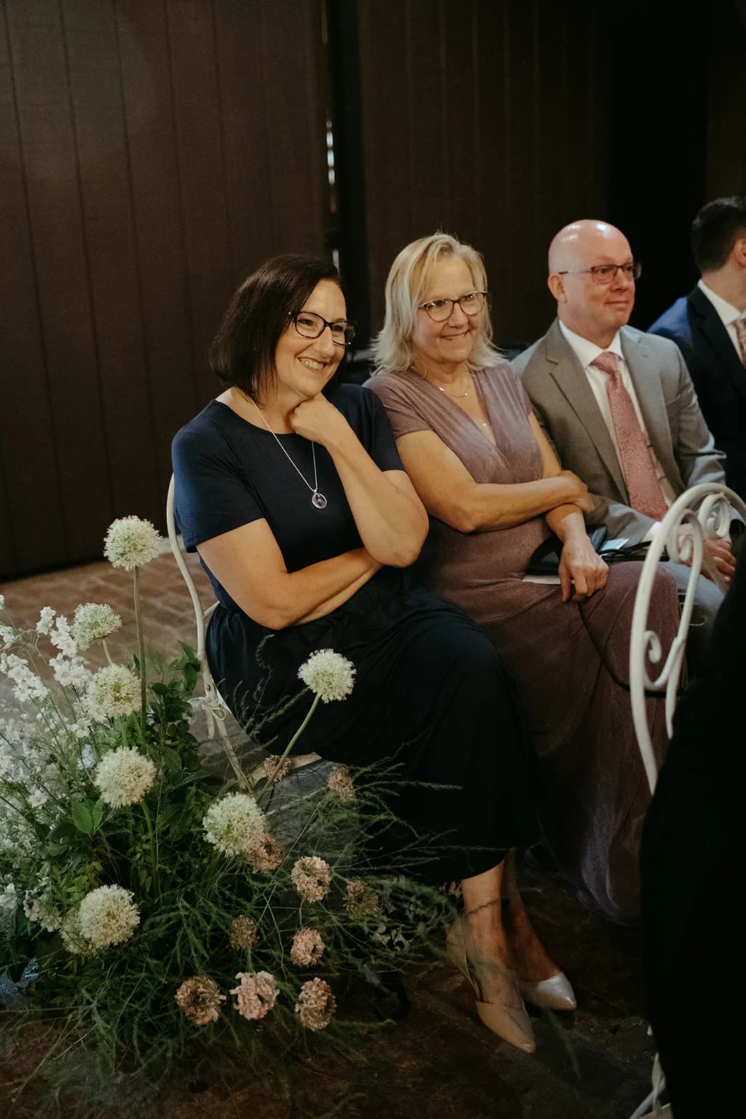 Three guests smile from white iron chairs beside an aisle arrangement of white allium, larkspur, scabiosa and fine grasses