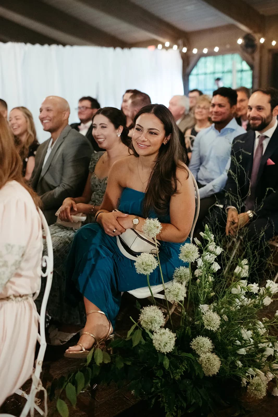 Guest in a teal gown watches the ceremony beside an aisle arrangement of white allium and campanula rising on tall stems