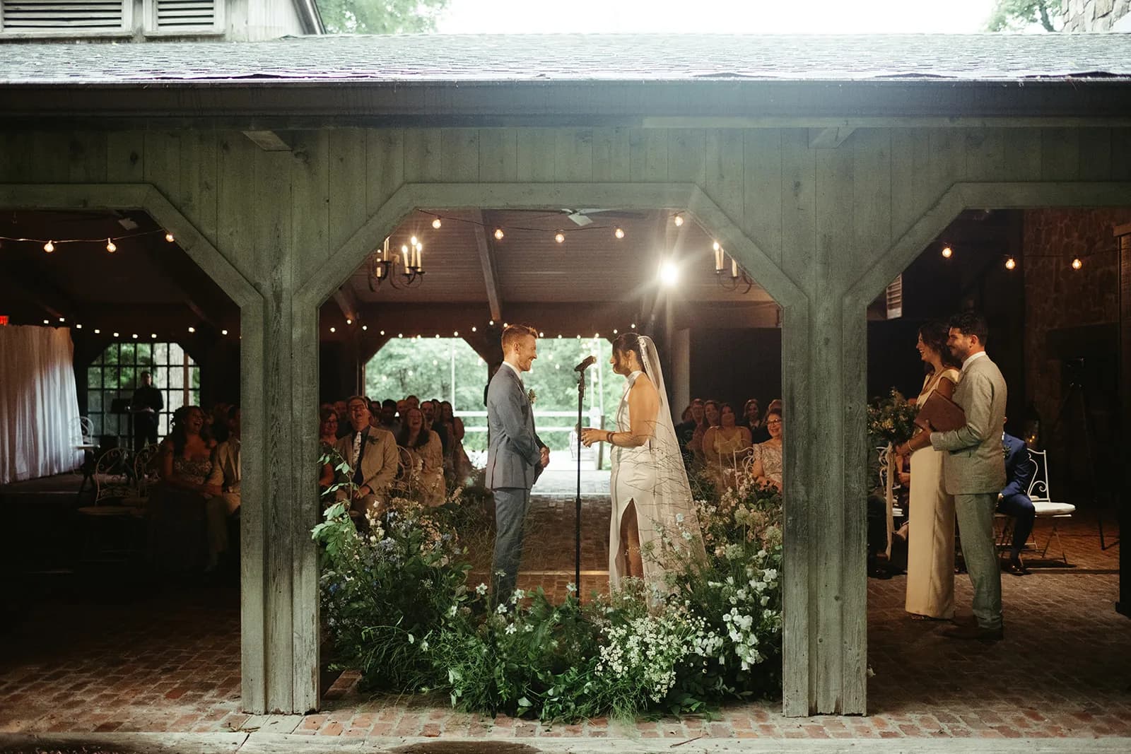 Through the pavilion posts, the couple stand at the microphone within a crescent of white and blue blooms as guests look on