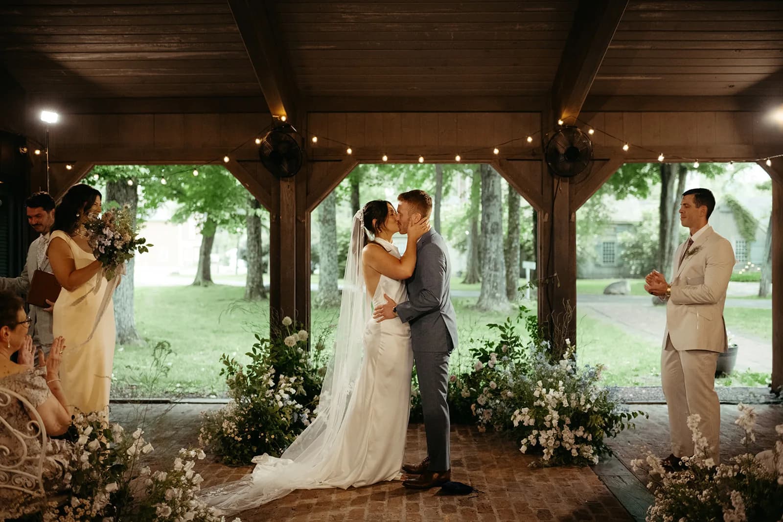 Shayna and Evan share a first kiss beneath the pavilion, campanula and delphinium massed along the brick floor behind them
