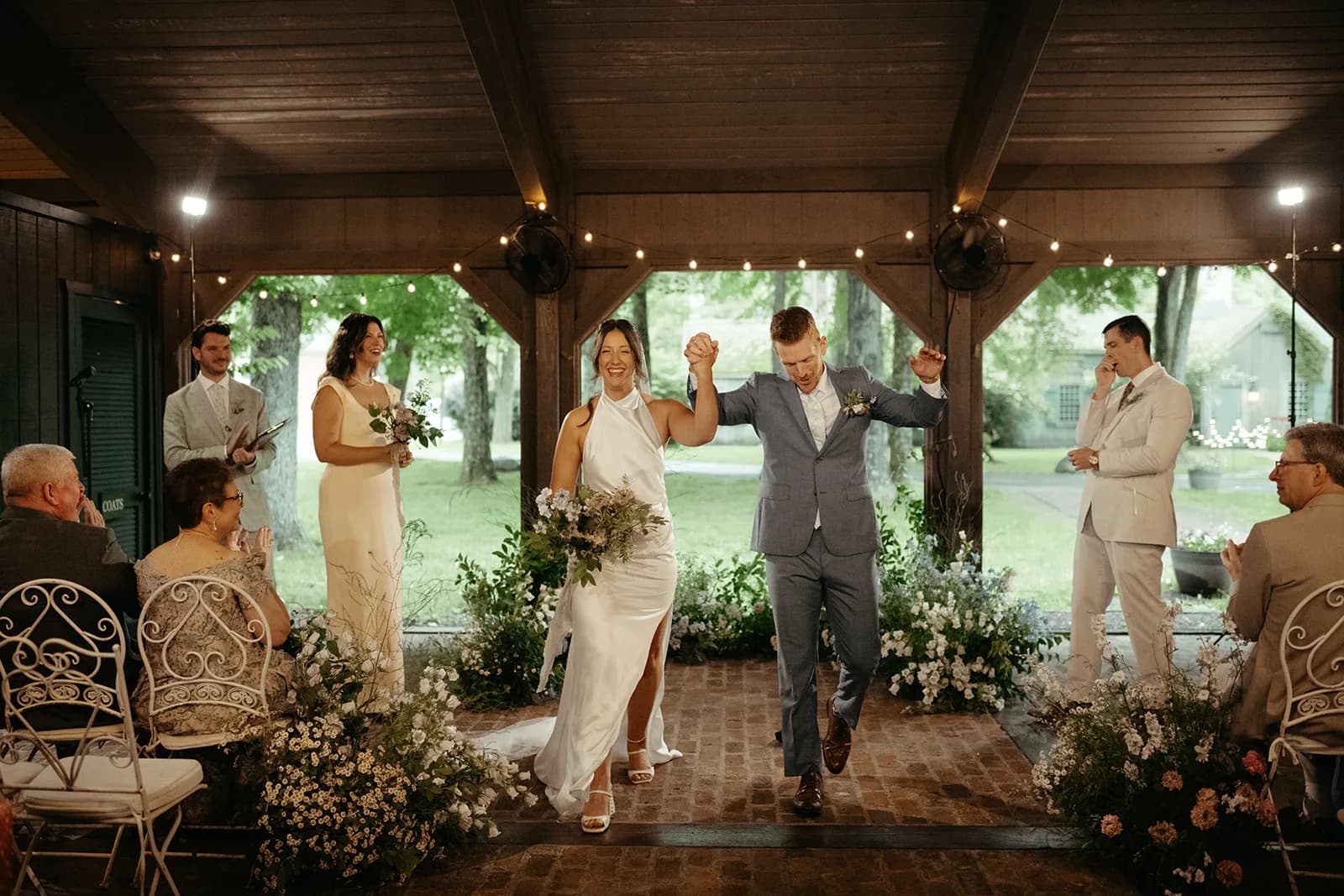 The newlyweds walk the brick aisle with joined hands raised, Shayna carrying her bouquet past banks of feverfew and campanula