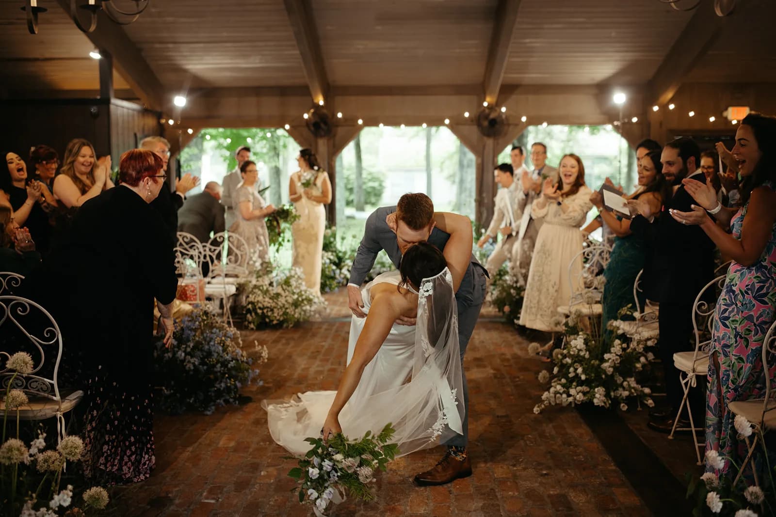 Evan dips Shayna in a kiss mid-aisle as guests applaud, her bouquet swinging low past white allium at the chair legs