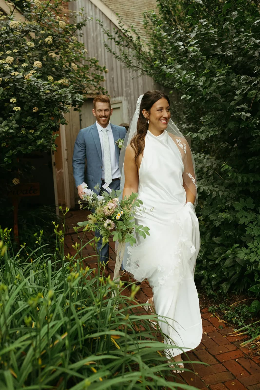 Shayna walks a brick garden path with a loose bouquet of scabiosa, feverfew and sweet pea, Evan following past viburnum