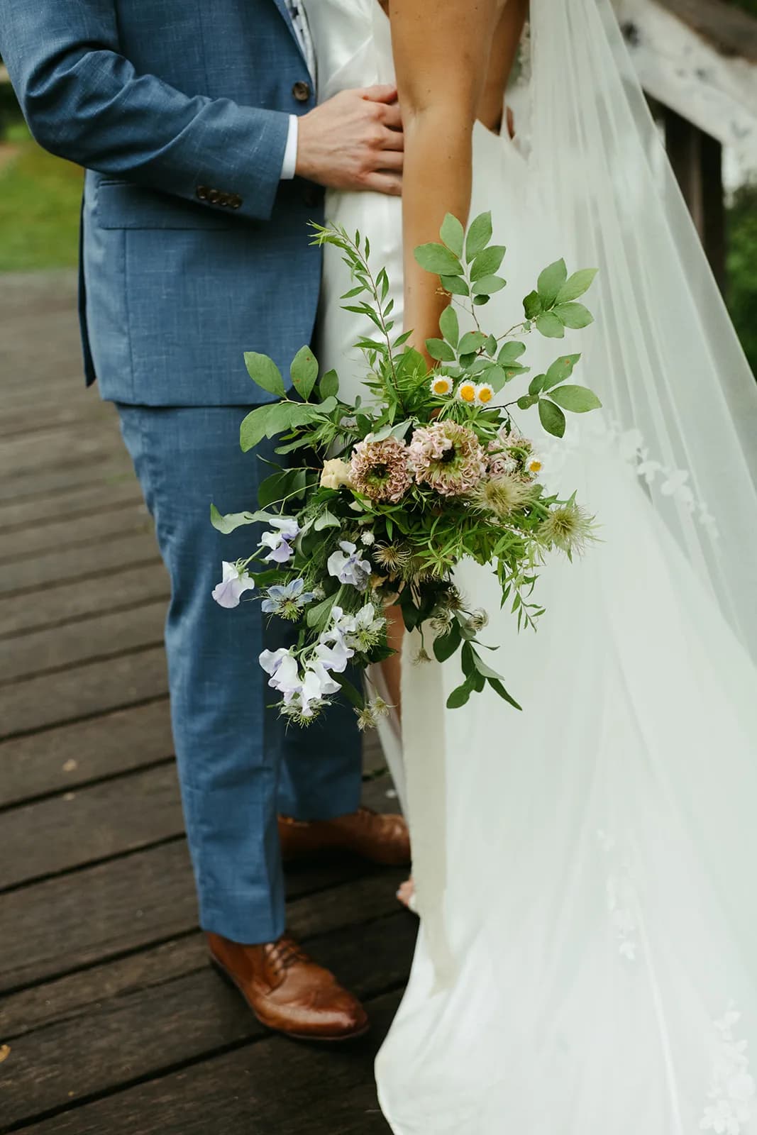 Blush scabiosa, pale blue sweet pea, nigella, feverfew and clematis seed heads trail from the bouquet held at Shayna's hip
