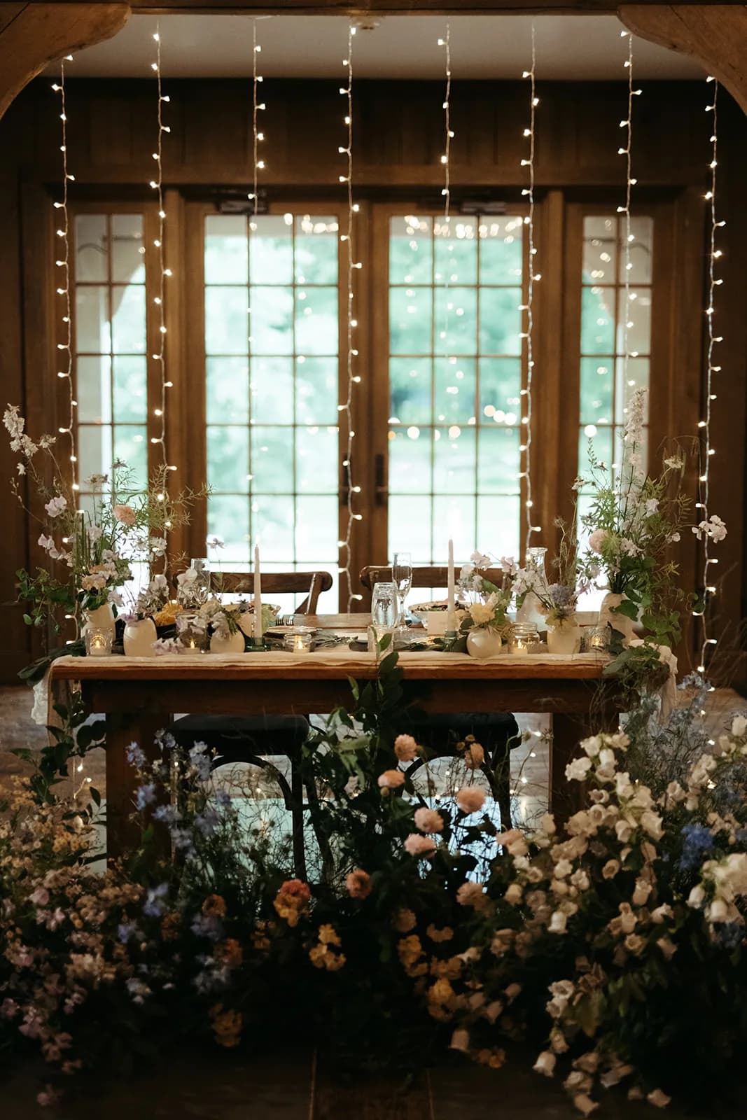 Sweetheart table before a curtain of string lights, asymmetric arrangements and tapers on the runner, blooms banked below