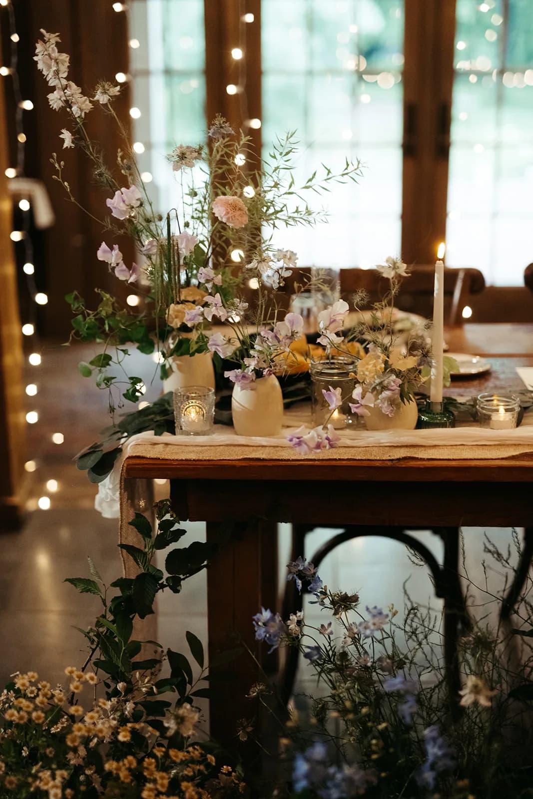 Asymmetric table arrangement of lilac sweet pea, larkspur, nigella and trailing ivy in cream vessels beside a taper on gauze