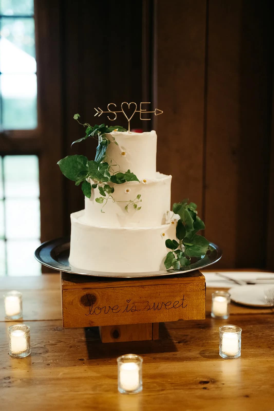 Three-tier white cake with trailing greenery and small daisies and a wire initials topper, set on a wooden box among votives
