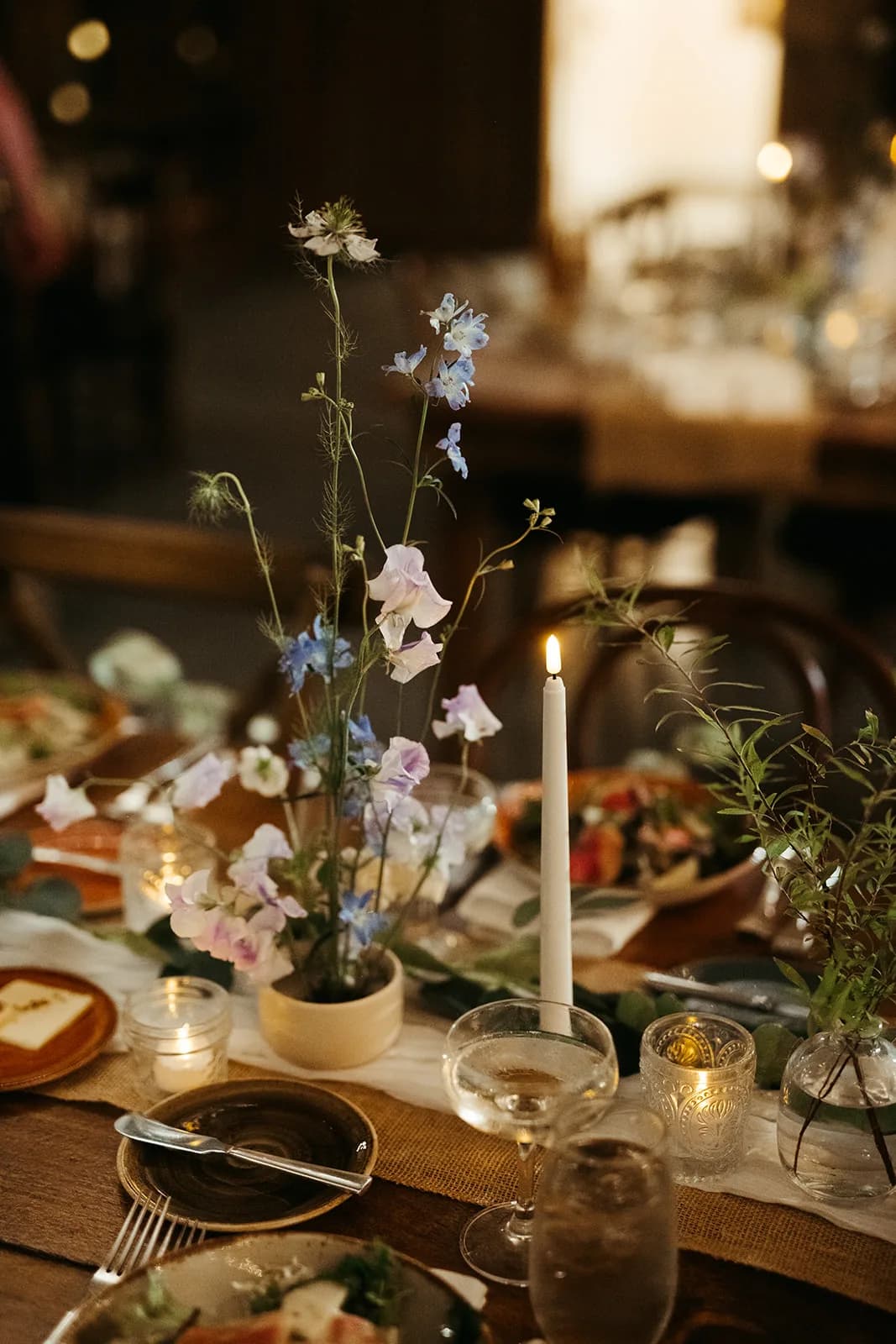 Tall nigella, spray delphinium and lilac sweet pea rise from a low ceramic vessel beside a taper candle and coupe glass