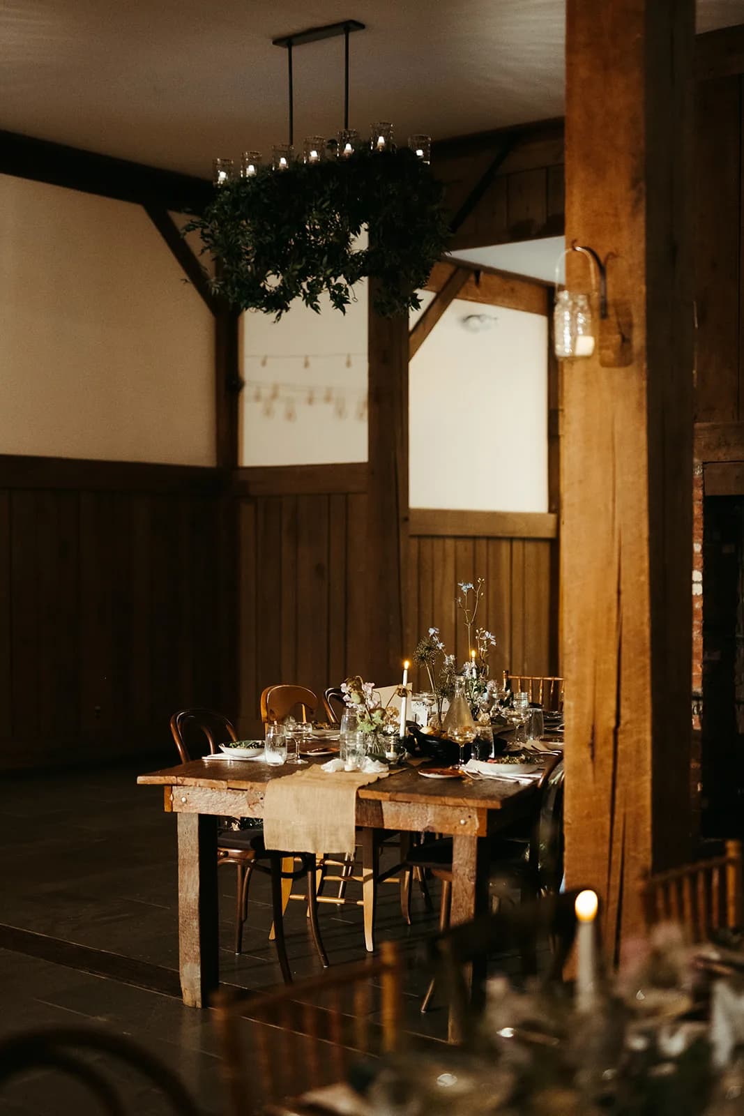 Long farm table under a greenery-wrapped candle chandelier, a burlap runner, sparse blue delphinium and tapers along it
