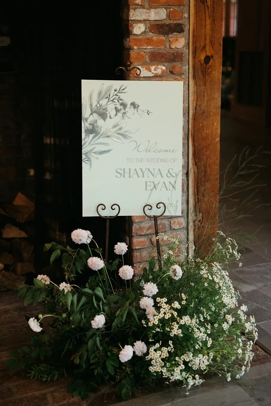 Welcome sign on an iron easel against a brick pillar, a ground arrangement of blush scabiosa, feverfew and foliage below