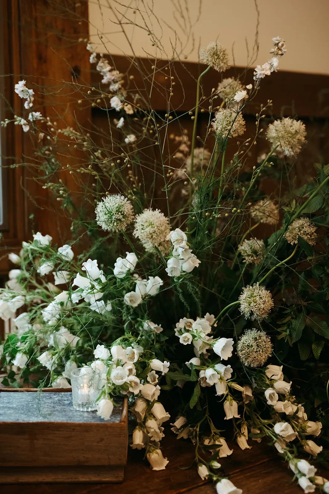 White allium, campanula and larkspur cascade over a zinc-topped crate with a votive, seeded stems rising against paneling