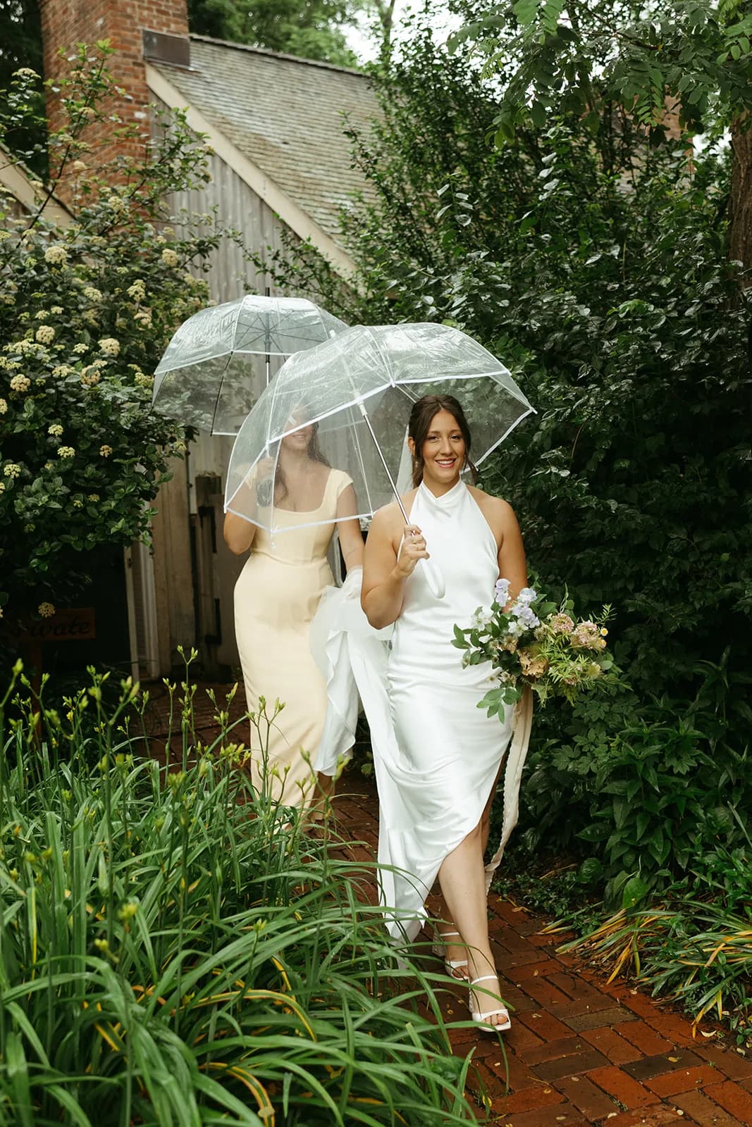 Shayna walks a brick path under a clear umbrella with a bouquet of sweet pea and scabiosa, an attendant in yellow following