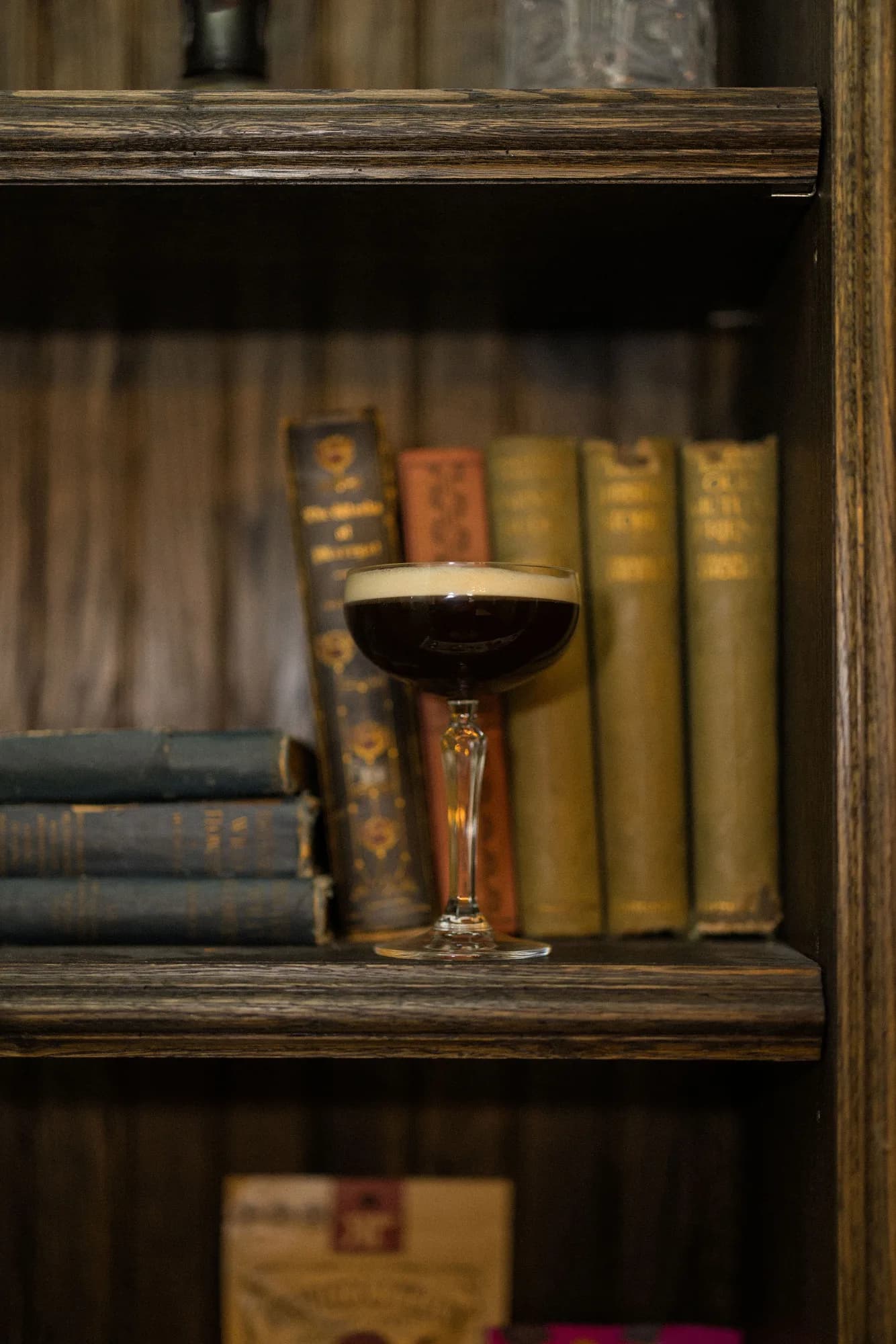 An espresso martini in a stemmed coupe rests on a dark oak library shelf among worn clothbound antique books