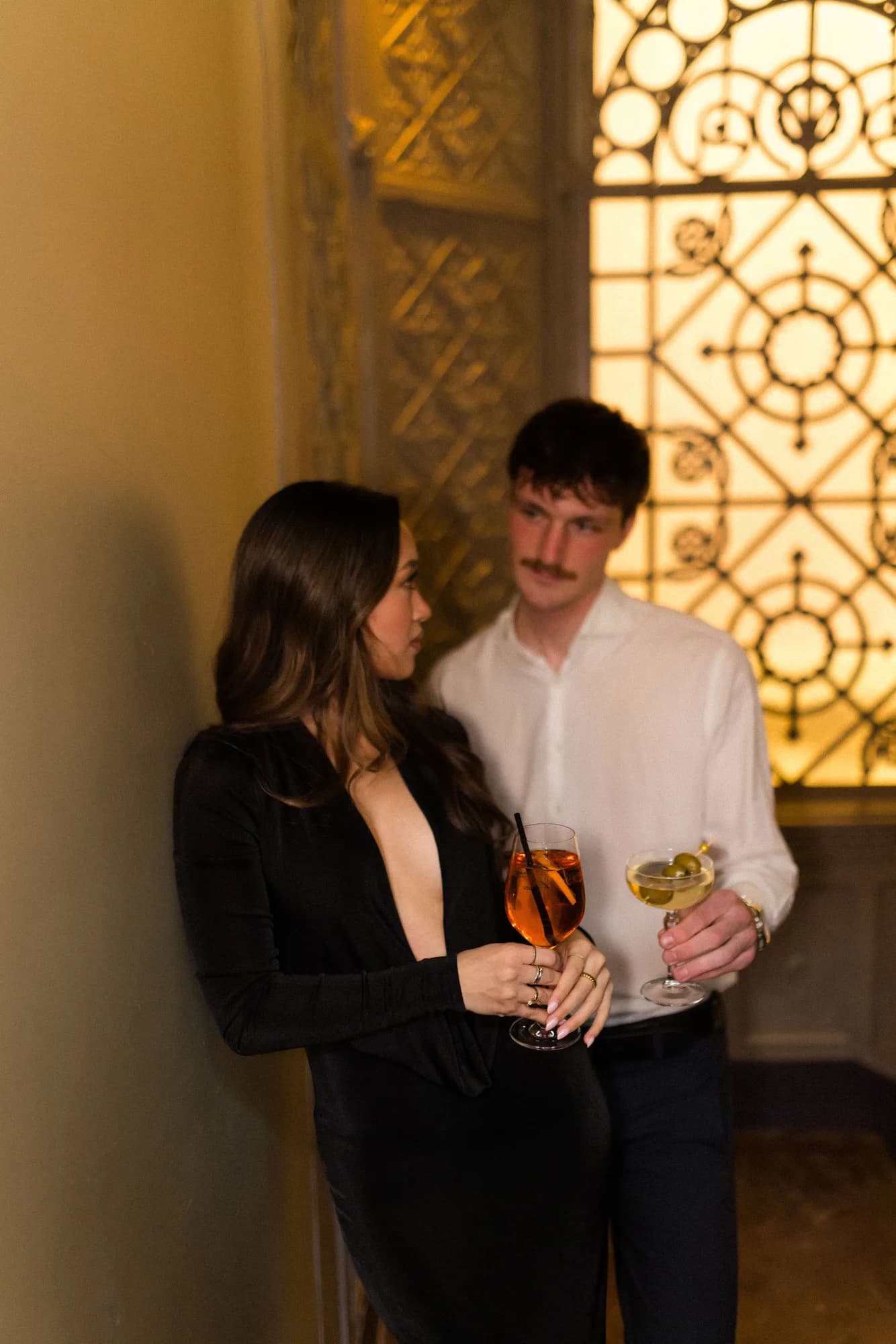 A couple in black and white leans close before a backlit ornamental iron screen, a spritz and a martini in hand