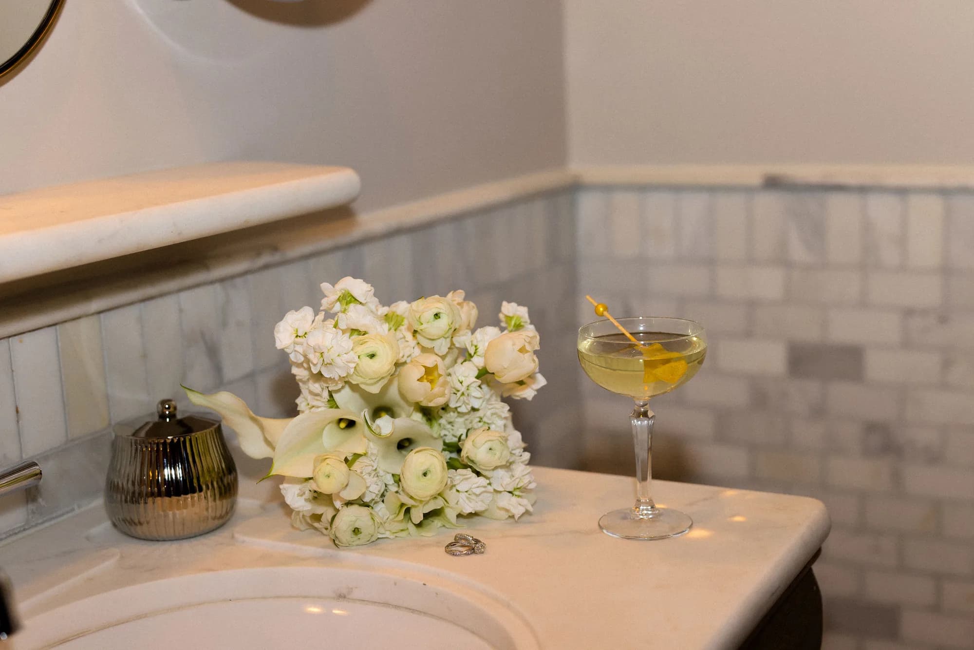 White ranunculus, tulips, stock and calla lilies rest on a marble vanity beside a lemon twist martini and two rings