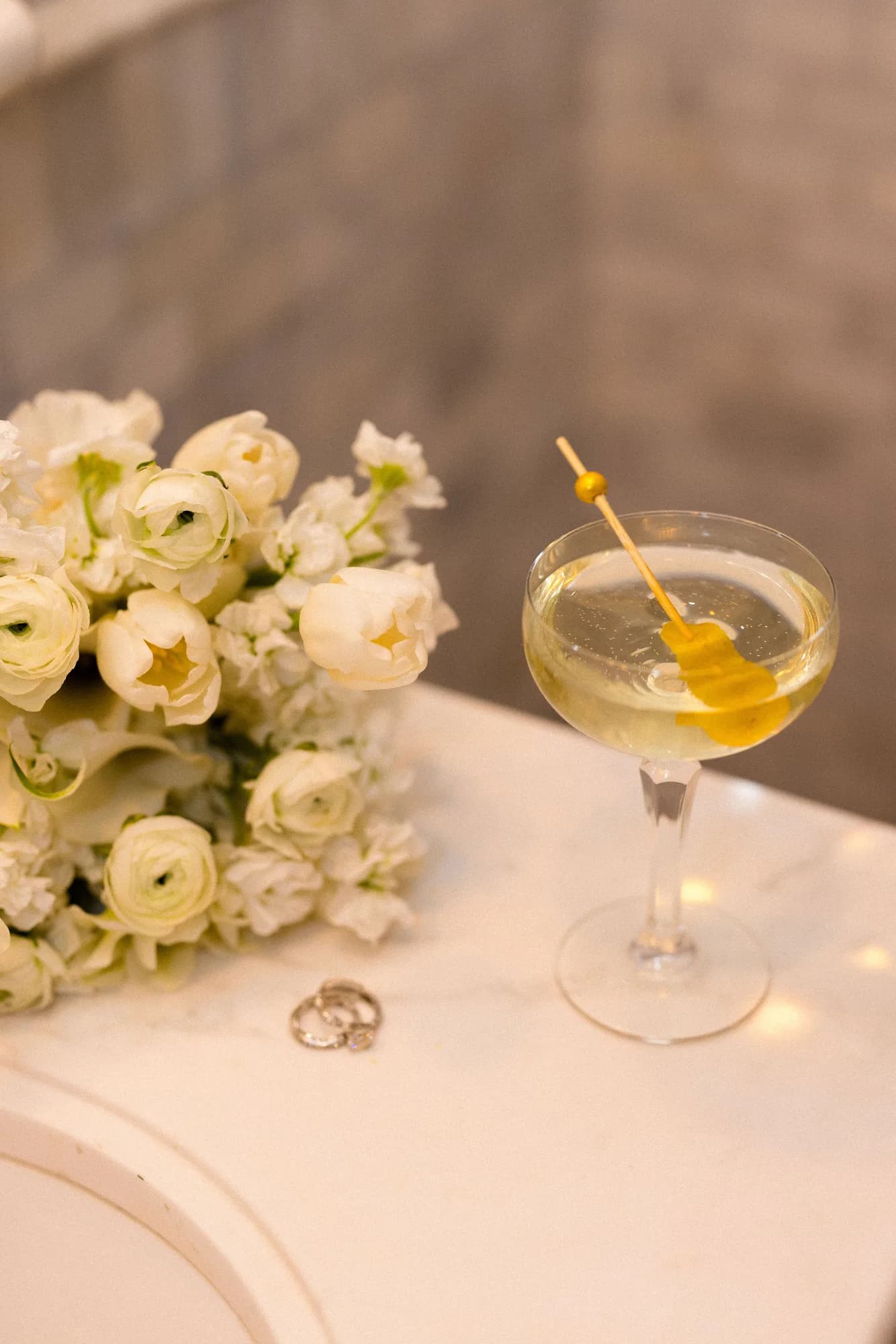 Close view of white tulips and ranunculus beside a coupe with a lemon twist on a gold pick, two rings on the marble