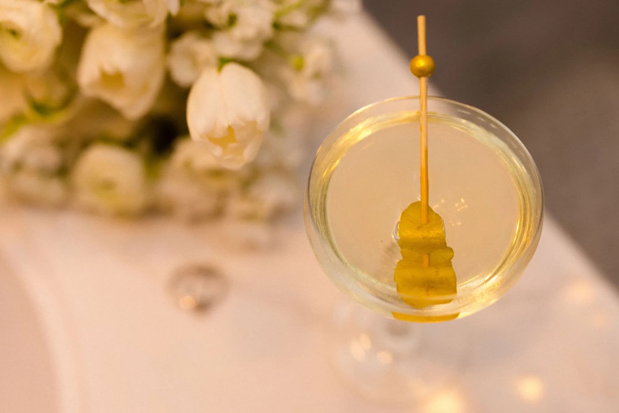 Overhead view of a martini coupe with a lemon twist on a gold-tipped pick, white tulips soft in the background