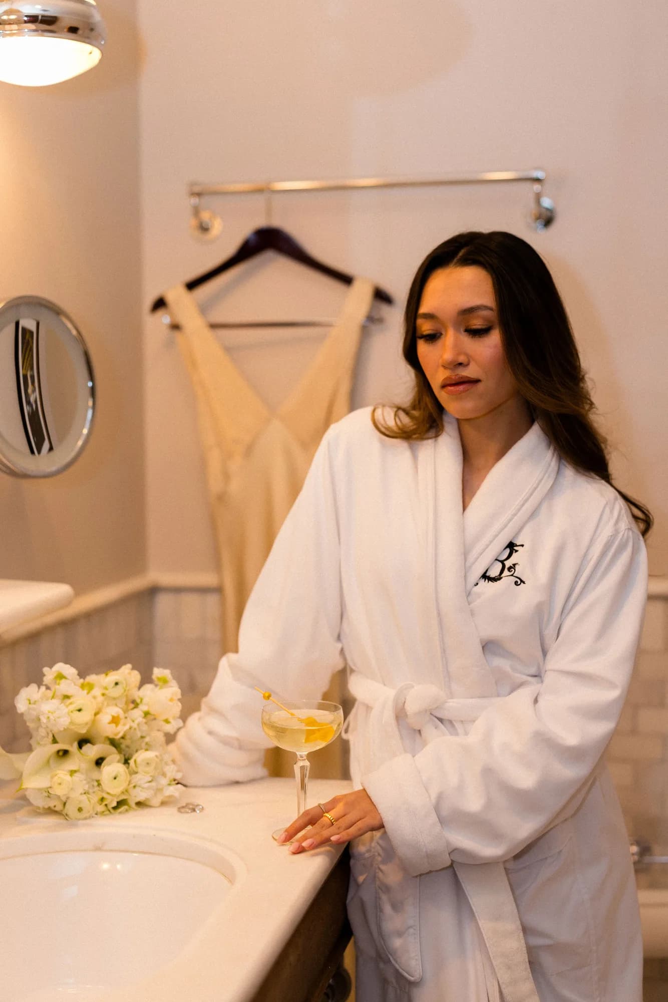 A woman in a white monogrammed robe pauses at a marble vanity with a white bouquet and a martini, a satin gown hanging behind
