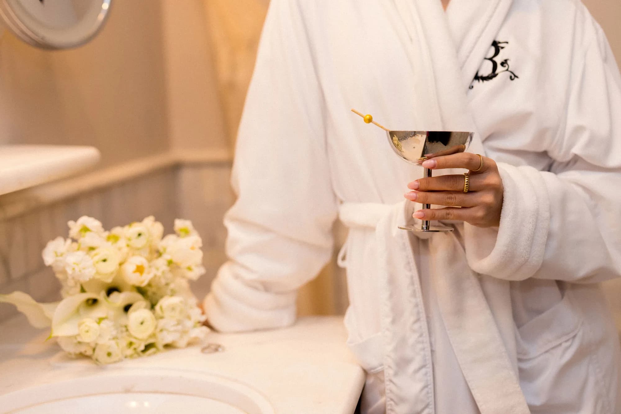 A hand in gold rings lifts a silver coupe above a marble vanity where a white bouquet of ranunculus and calla lilies rests