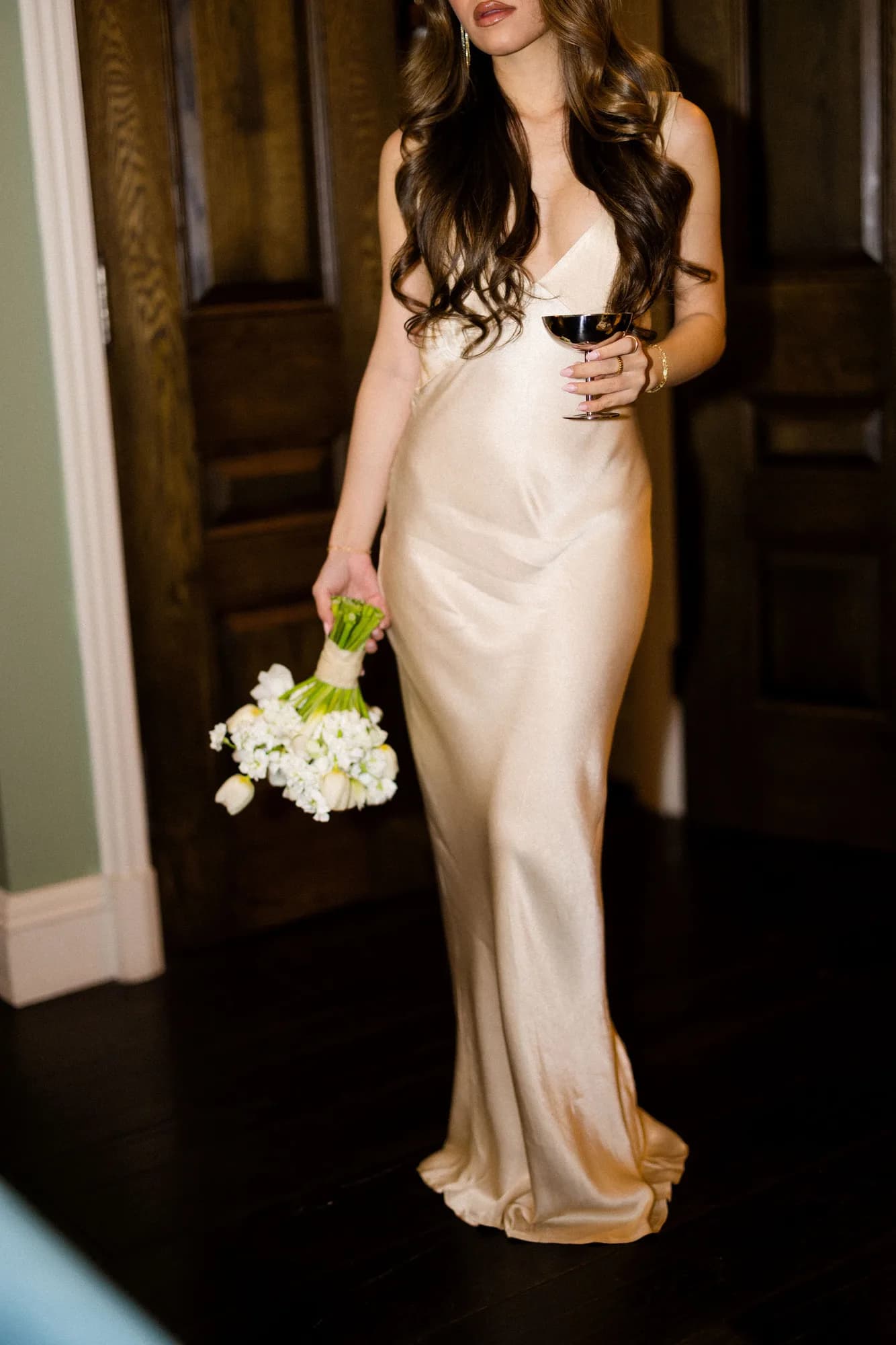 A bride in a champagne satin slip gown carries a ribbon-wrapped white bouquet at her side and a silver coupe before oak doors