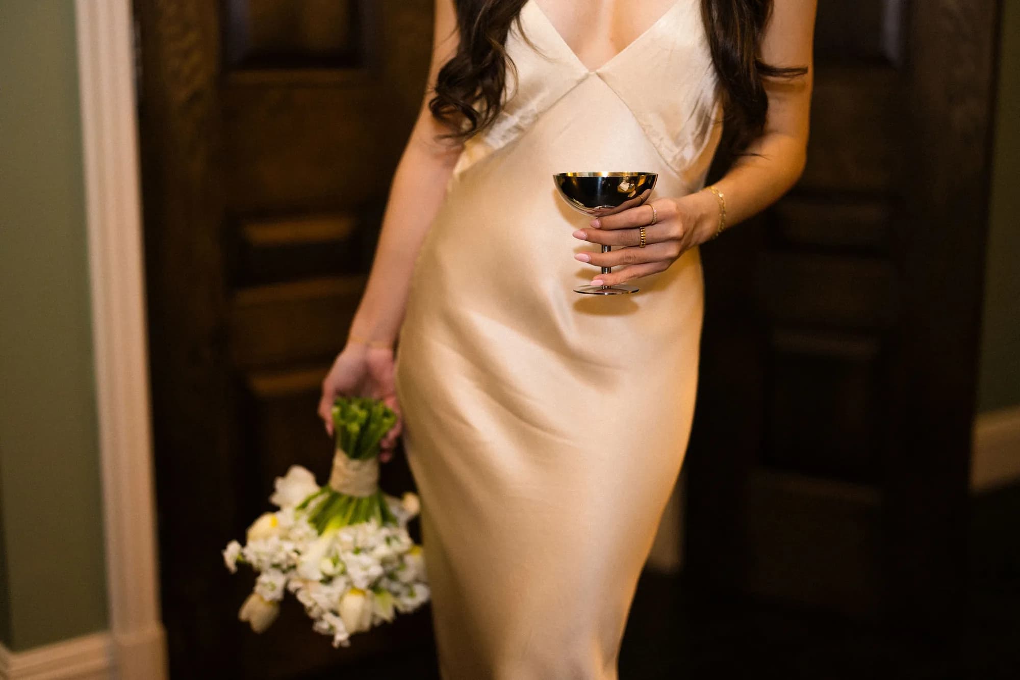 Close crop of a satin slip gown, a silver coupe held at the waist and a white bouquet with wrapped stems carried low